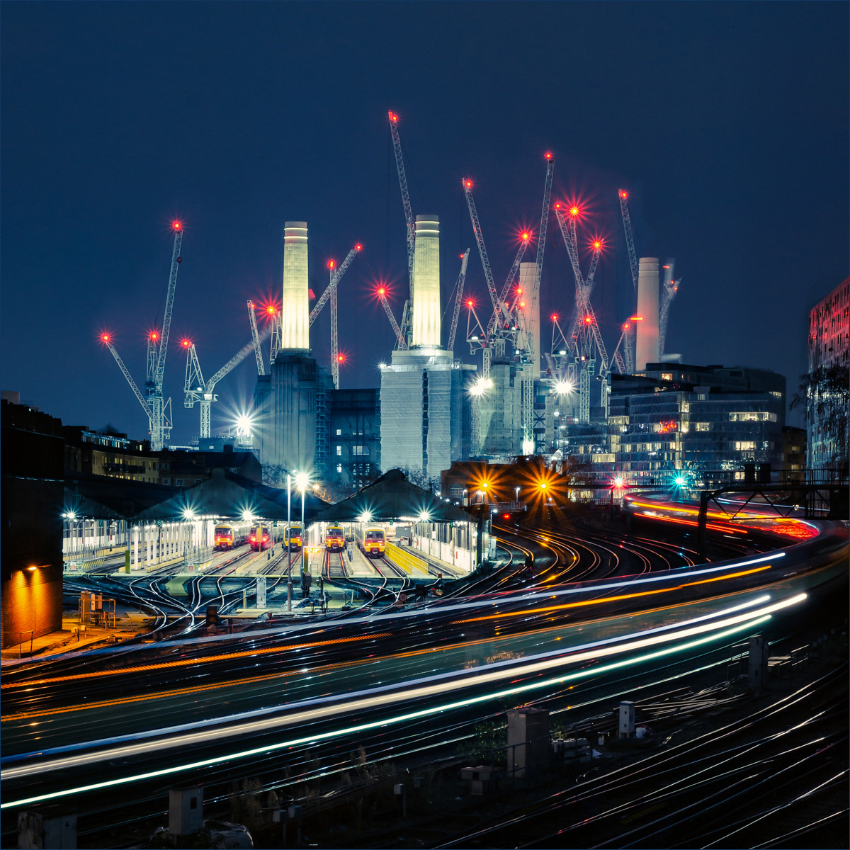 This dynamic night shot captures the iconic Battersea Power Station in London, surrounded by a forest of construction cranes. The illuminated chimneys stand tall against the dark sky, while red crane lights dot the scene like stars. In the foreground, the train tracks and platforms are bustling with activity, with multiple trains parked and light trails indicating the movement of passing trains. The long exposure effect enhances the sense of motion and energy, highlighting the transformation of this historic site amidst ongoing redevelopment.