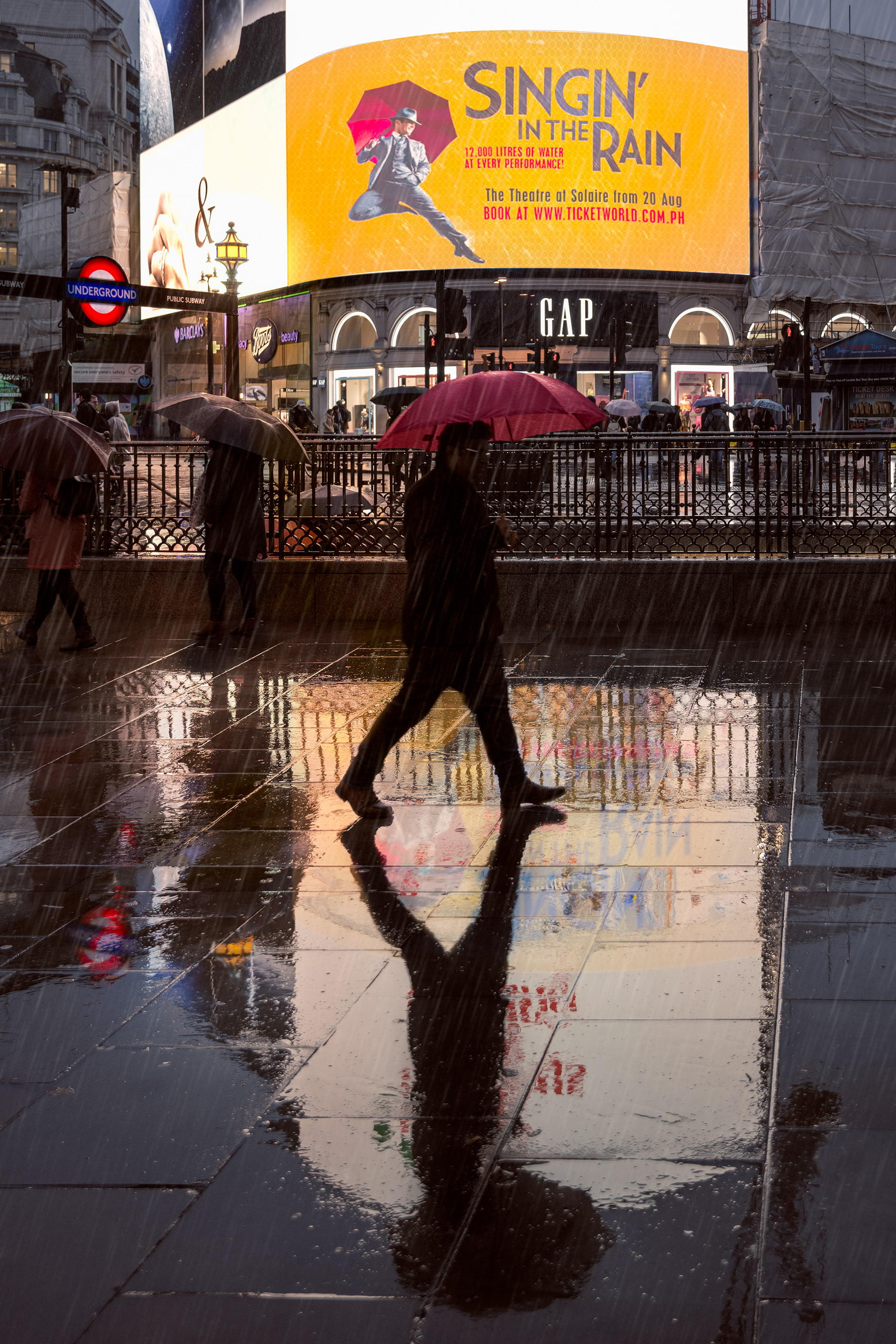A wet scene in Piccadilly Circus, London. A person holding a red umbrella walks through a rain-soaked plaza in front of an illuminated billboard advertising "Singin' in the Rain." The scene is bustling with people carrying umbrellas, and the wet ground reflects the bright lights of the advertisement and surrounding buildings. The vibrant colors and reflections create a lively atmosphere despite the rainy weather.
