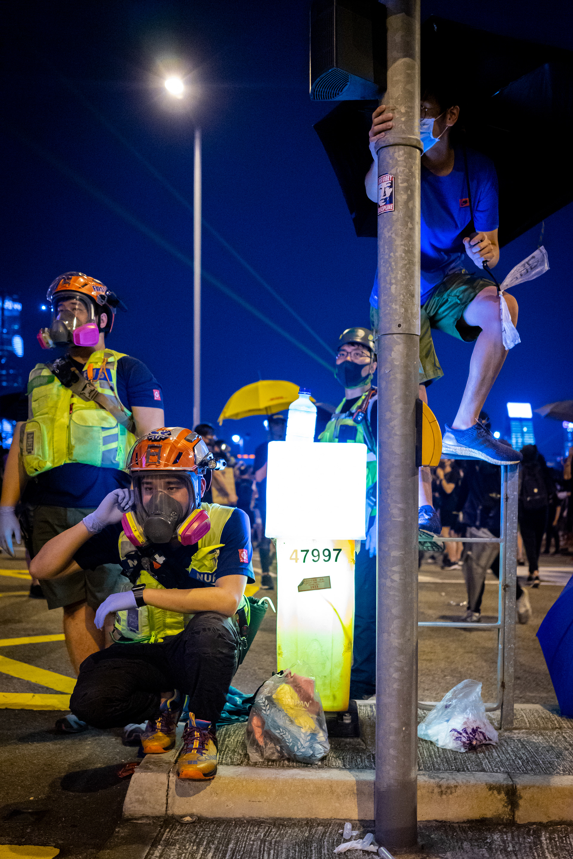 The Wait: Admiralty, Hong Kong: Nok, the leader of a volunteer EMS team waits with his crew while police douse citizens with a fire hose. Citizens gathered at Tamar Park to commemorate the fifth anniversary of the Umbrella Movement protests. September 28, 2019-Hong Kong