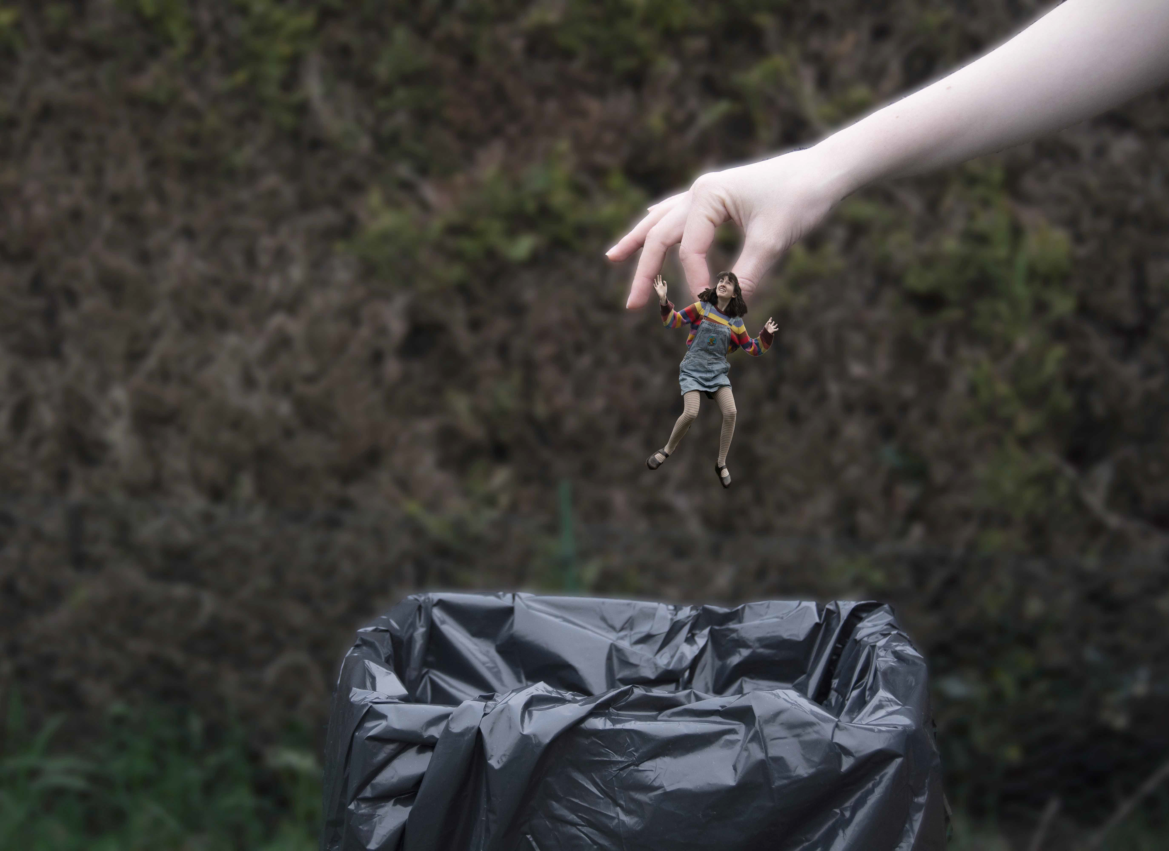 A woman is picked-up by a large hand and placed into a wastebin.