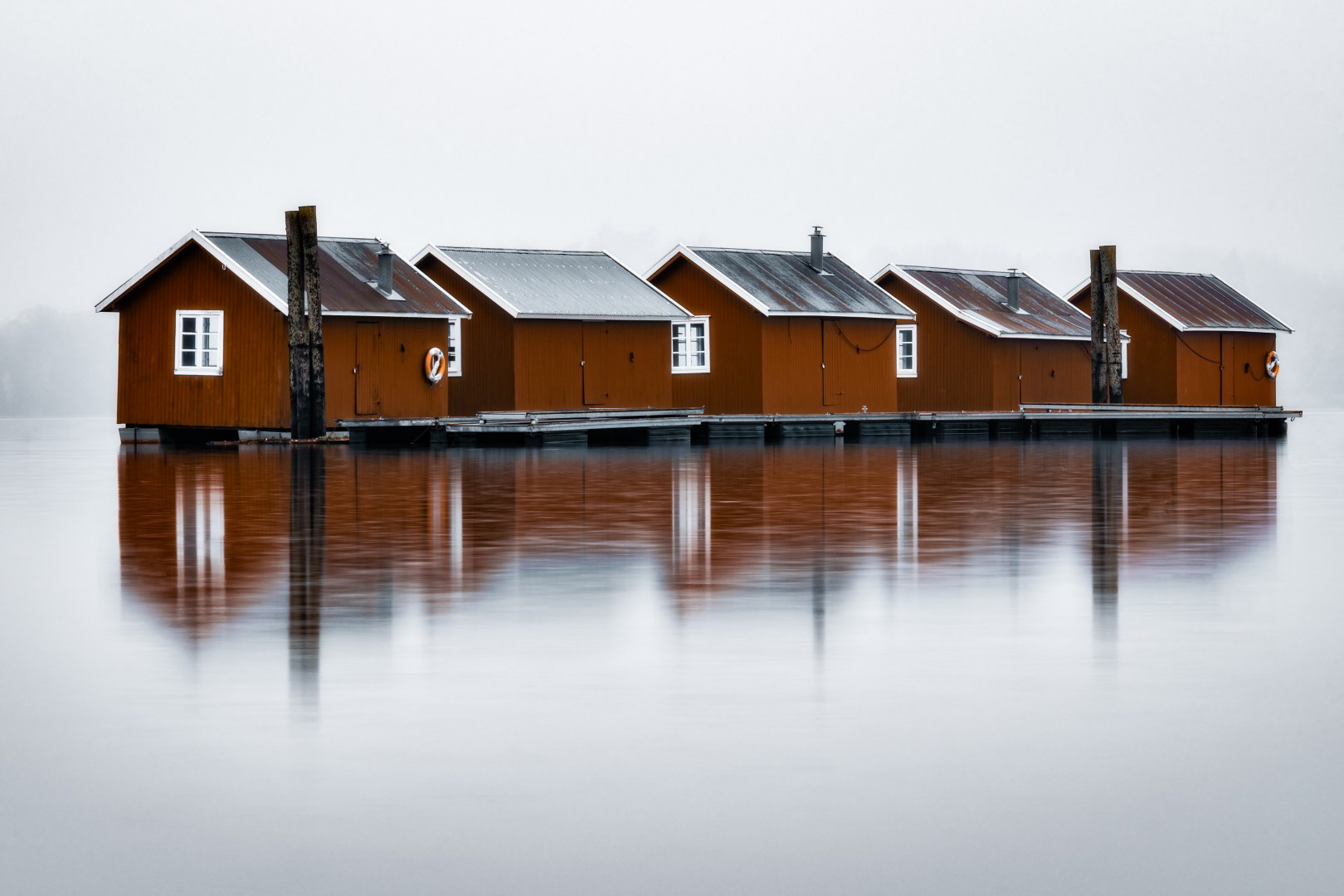 5 røde pausebrakker flytende i Glomma. Tåke med duse rødfarger.