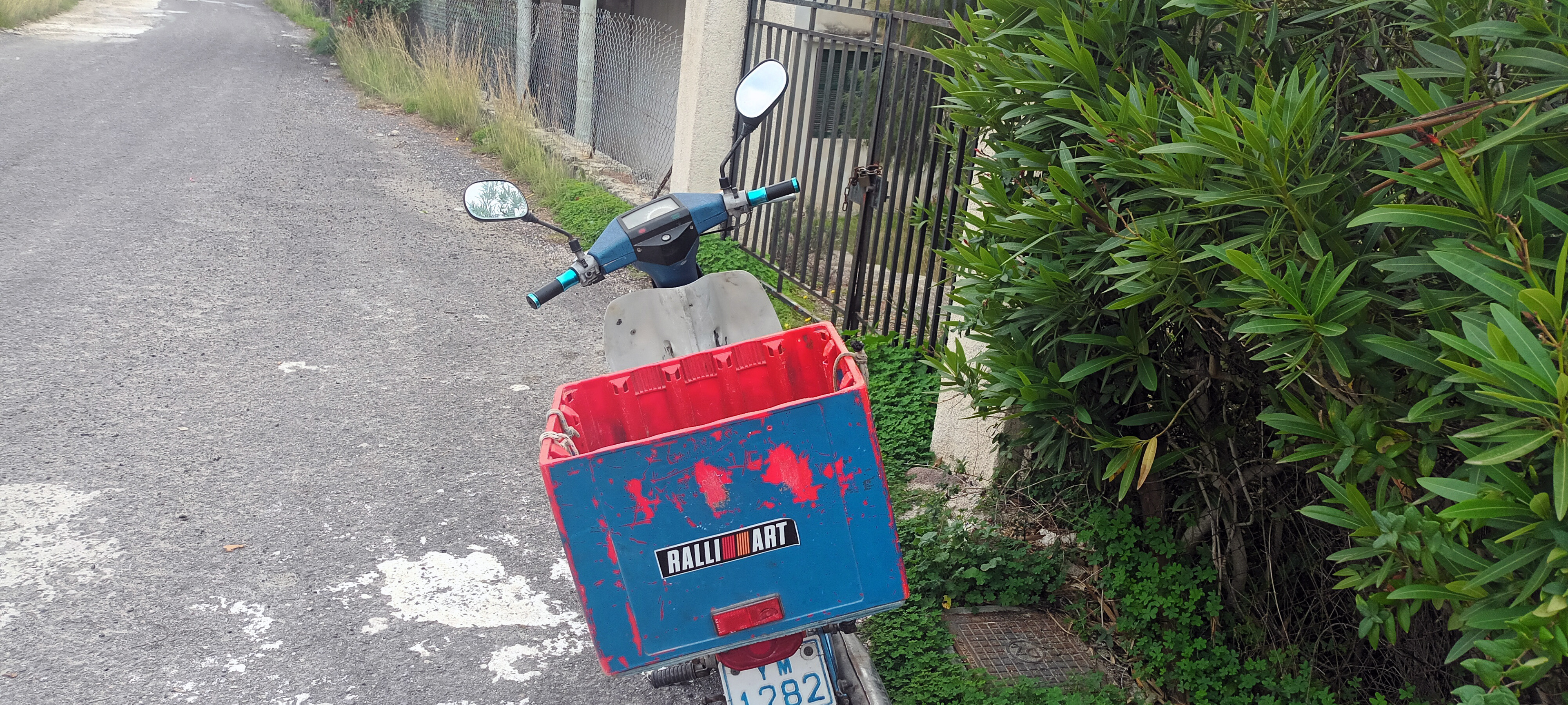 An old YAMAHA V50 moped with Ένα παλιό παπί YAMAHA V50 με μπλε-κόκκινο καφάσι και αυτοκόλλητο "Ralli Art" στέκεται σε στενό δρομάκι της Αίγινας.a blue-red crate and a "Ralli Art" sticker stands in a narrow Aegina street.