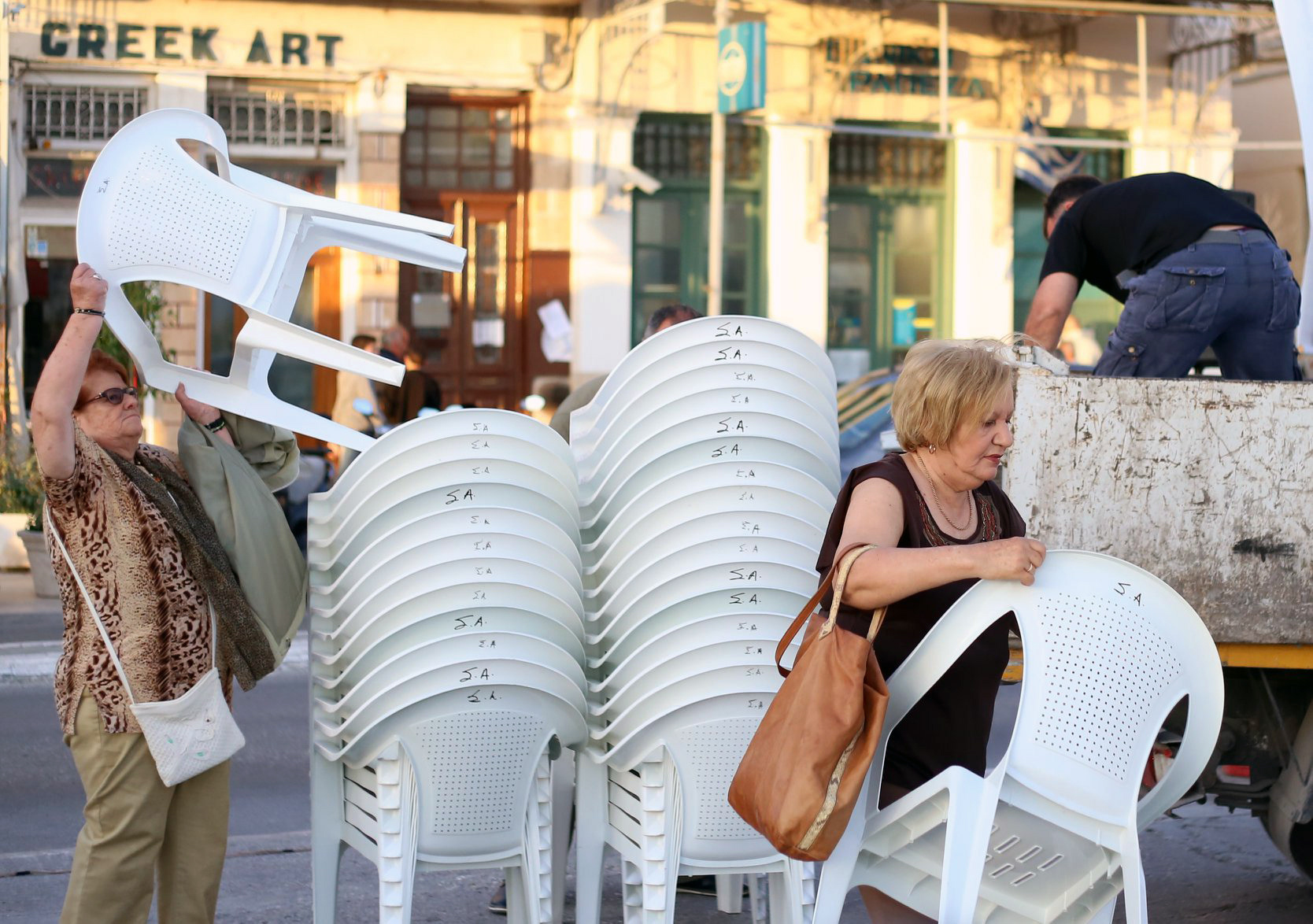Women organizing and carrying stacked white Monobloc chairs in a busy urban setting, showcasing their practicality and cultural presence.Γυναίκες οργανώνουν και μεταφέρουν στοιβαγμένες λευκές καρέκλες Monobloc σε ένα πολυσύχναστο αστικό περιβάλλον, αναδεικνύοντας την πρακτικότητα και την πολιτισμική τους παρουσία.