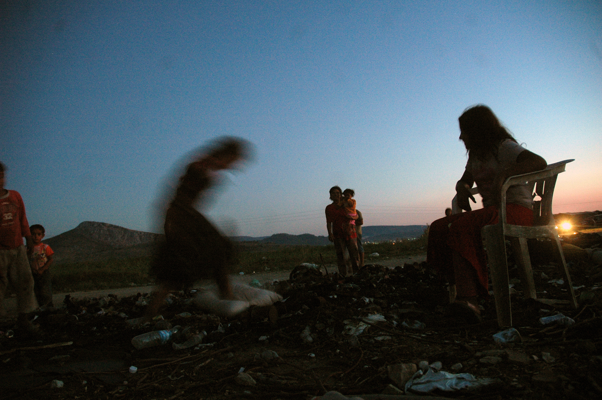 Silhouette of a woman on a white plastic monobloc chair amidst trash, with children in the background and the horizon at sunset.Σιλουέτα γυναίκας σε λευκή πλαστική καρέκλα ανάμεσα σε σκουπίδια, με παιδιά στο βάθος και τον ορίζοντα στο ηλιοβασίλεμα