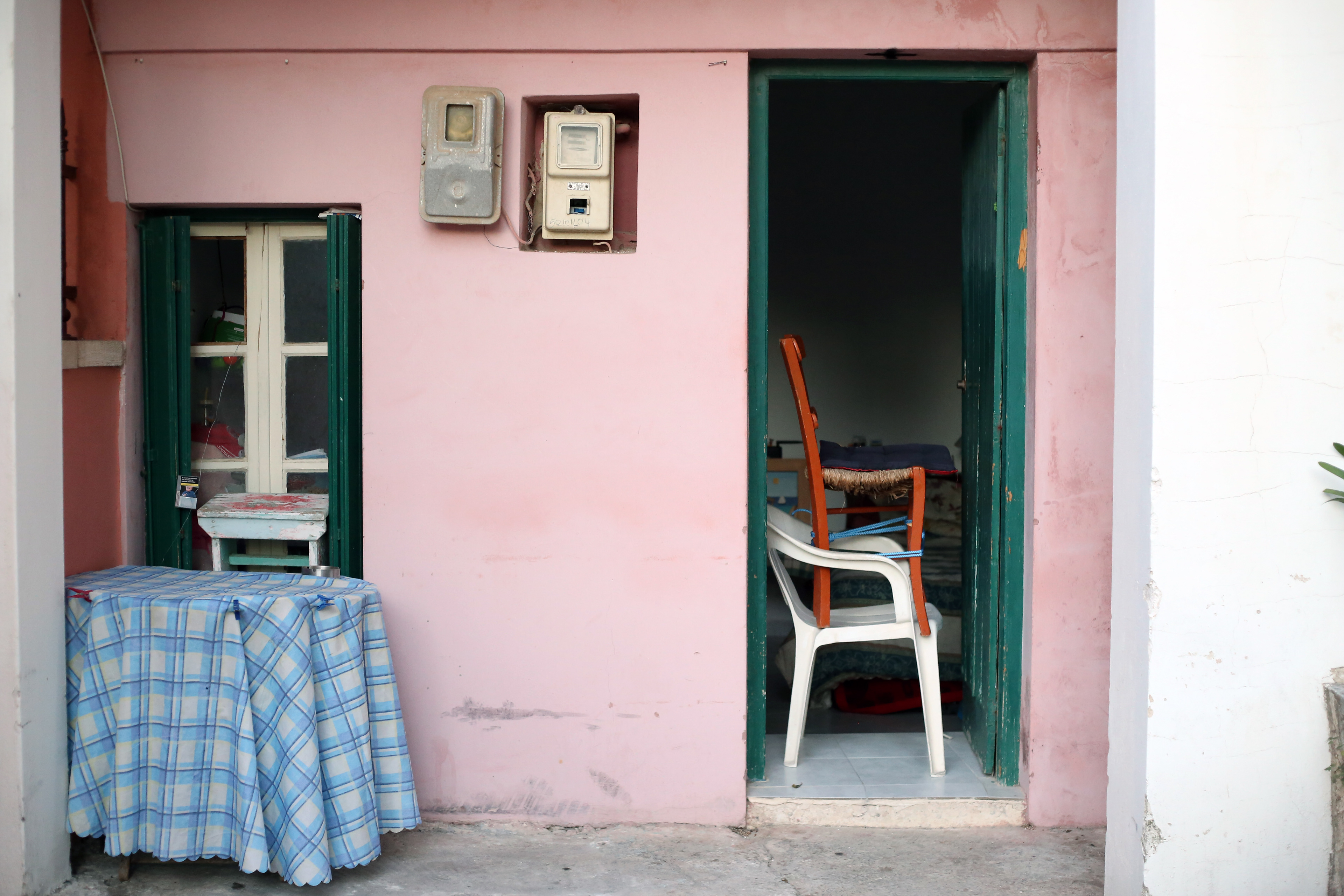 White plastic chair stacked with a wooden chair in a doorway, surrounded by a pink wall, a table with a blue cloth, and a window.White plastic chair stacked with a wooden chair in a doorway, surrounded by a pink wall, a table with a blue cloth, and a window.