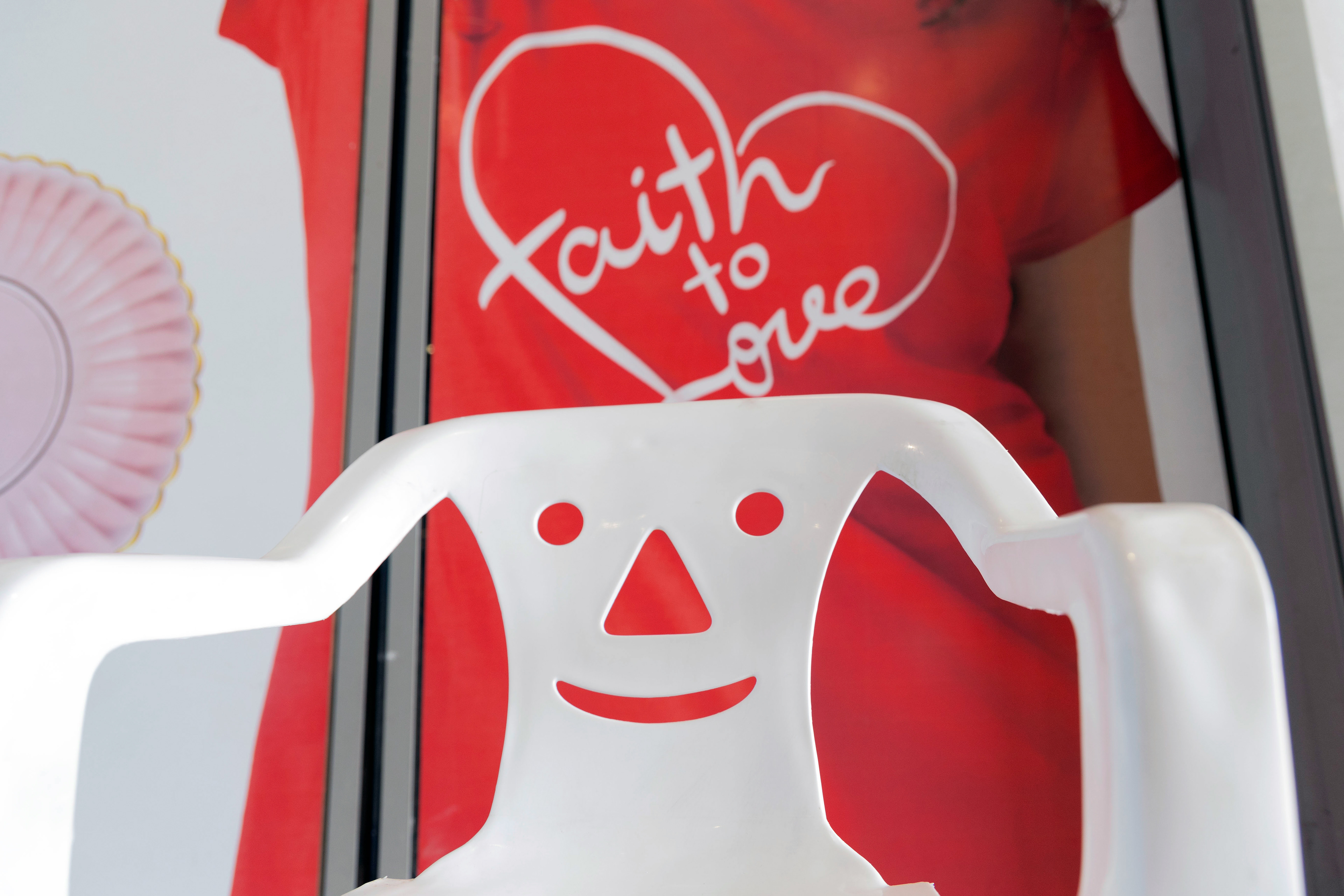 White Monobloc chair with a smiley face design, set against a red backdrop with 'Faith to Love' text, symbolizing warmth and optimism.Λευκή καρέκλα Monobloc με σχέδιο χαμογελαστού προσώπου, τοποθετημένη σε κόκκινο φόντο με το κείμενο "Faith to Love", συμβολίζοντας τη ζεστασιά και την αισιοδοξία.