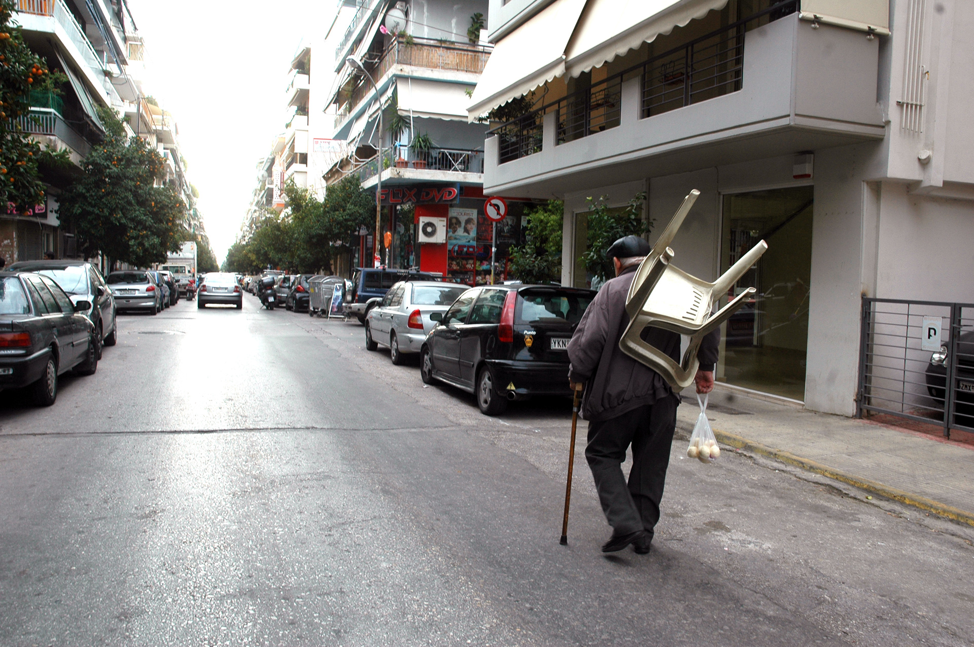 An elderly man walks down an urban street with a white plastic monobloc chair on his back, a cane in one hand, and a bag of eggs in the other.Ένας ηλικιωμένος άνδρας περπατά σε έναν αστικό δρόμο με μια λευκή πλαστική καρέκλα στην πλάτη του, ένα μπαστούνι στο ένα χέρι και μια σακούλα με αυγά στο άλλο