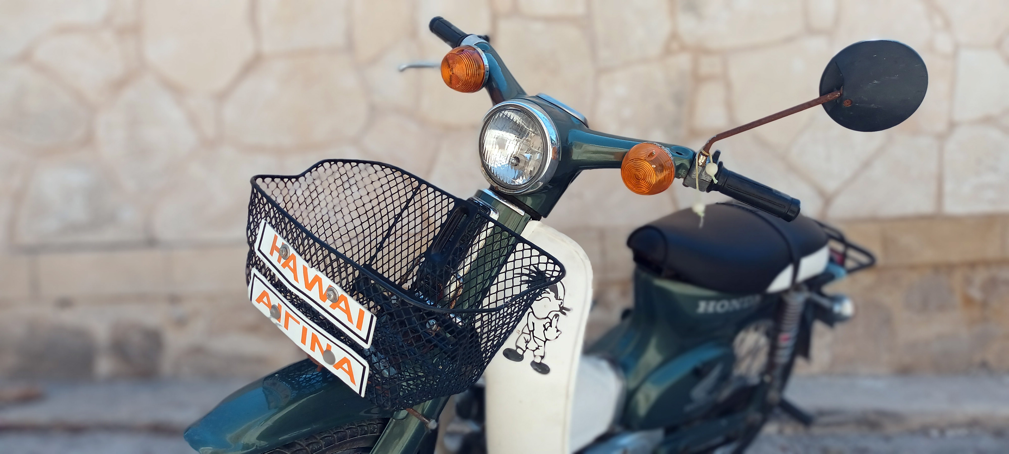 A green Honda C50 moped with a basket labeled "HAWAII AIGINA," a rusty mirror, and custom decorations, leaning against a stone wall.Ένα πράσινο Honda C50 παπί με καλάθι που γράφει "HAWAII ΑΙΓΙΝΑ", σκουριασμένο καθρέφτη και αυτοσχέδια διακόσμηση, ακουμπά σε πέτρινο τοίχο.
