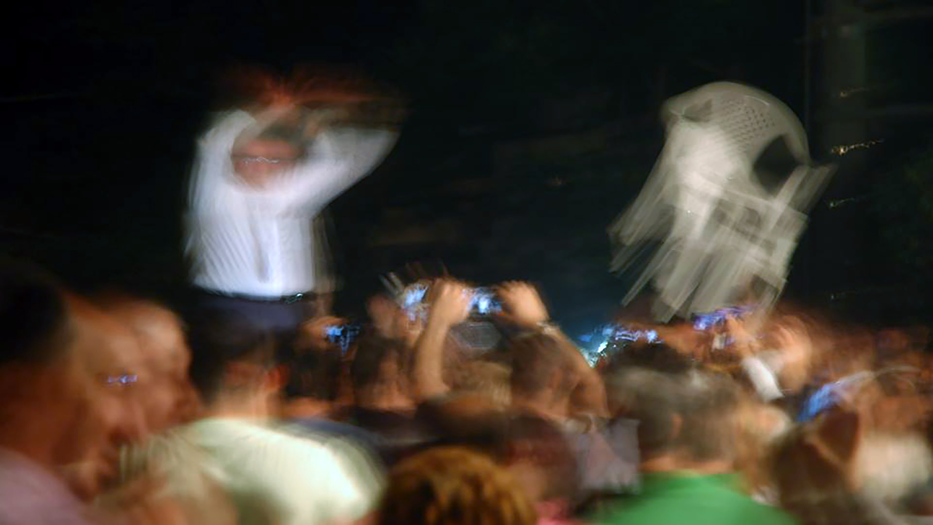 A motion-blurred scene of a white plastic monobloc chair thrown into a lively crowd, capturing its symbolic cultural role.Μια σκηνή με θολή κίνηση, όπου μια λευκή πλαστική καρέκλα monobloc πετιέται μέσα σε ένα ζωηρό πλήθος, αποτυπώνοντας τον συμβολικό της πολιτισμικό ρόλο.