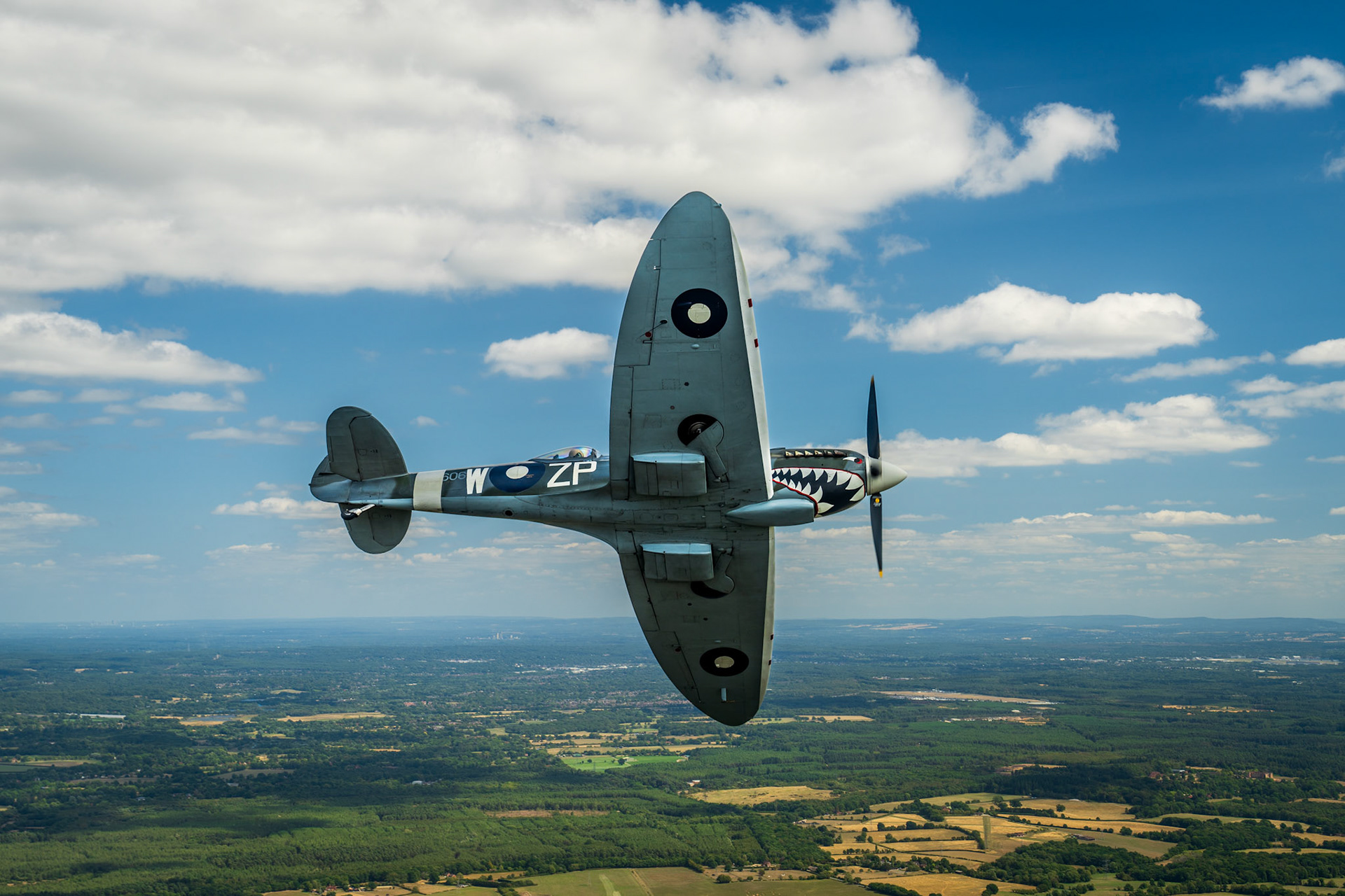 A Supermarine Spitfire in RAF markings — codes ZP-W and a shark-mouth nose detail — banks steeply above a patchwork of summer fields and woodland. The air-to-air perspective places the aircraft centre-frame against a broken cumulus sky, the rolling English lowlands receding to a distant horizon beneath it.