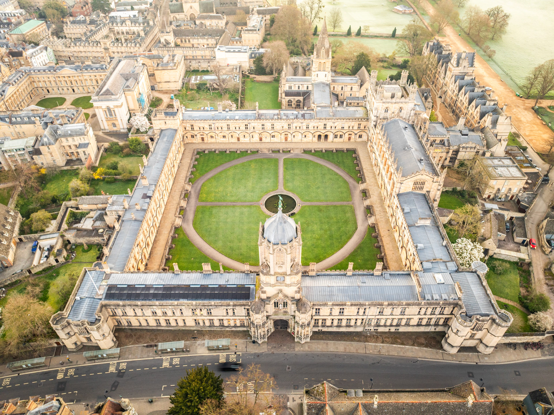 The symmetrical courtyard of All Souls College, Oxford, resolves from directly overhead into a geometry of limestone colonnades and circular lawn paths centred on a small fountain. Shot in flat spring light on 22 March, the overcast sky strips the scene of shadow drama but renders the Cotswold stone evenly, emphasising pattern over atmosphere. The image is competent and well-framed but reads as architectural documentation rather than a distinctive aerial landscape.