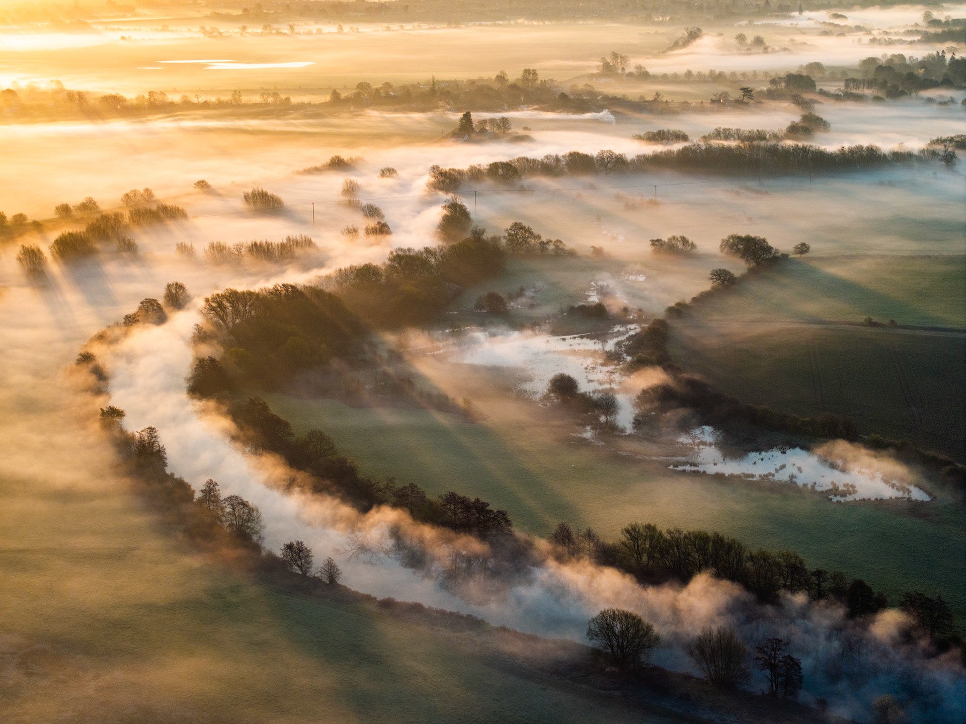 A meandering river valley near Wallingford is submerged beneath a temperature inversion at dawn, the mist pooling along the watercourse and tracing every curve of the floodplain. Bare riparian trees emerge as silhouettes through the vapour while crepuscular rays fan across the scene from low in the west. Flooded fields catch the light between the fog banks, giving the aerial view an almost topographic clarity.