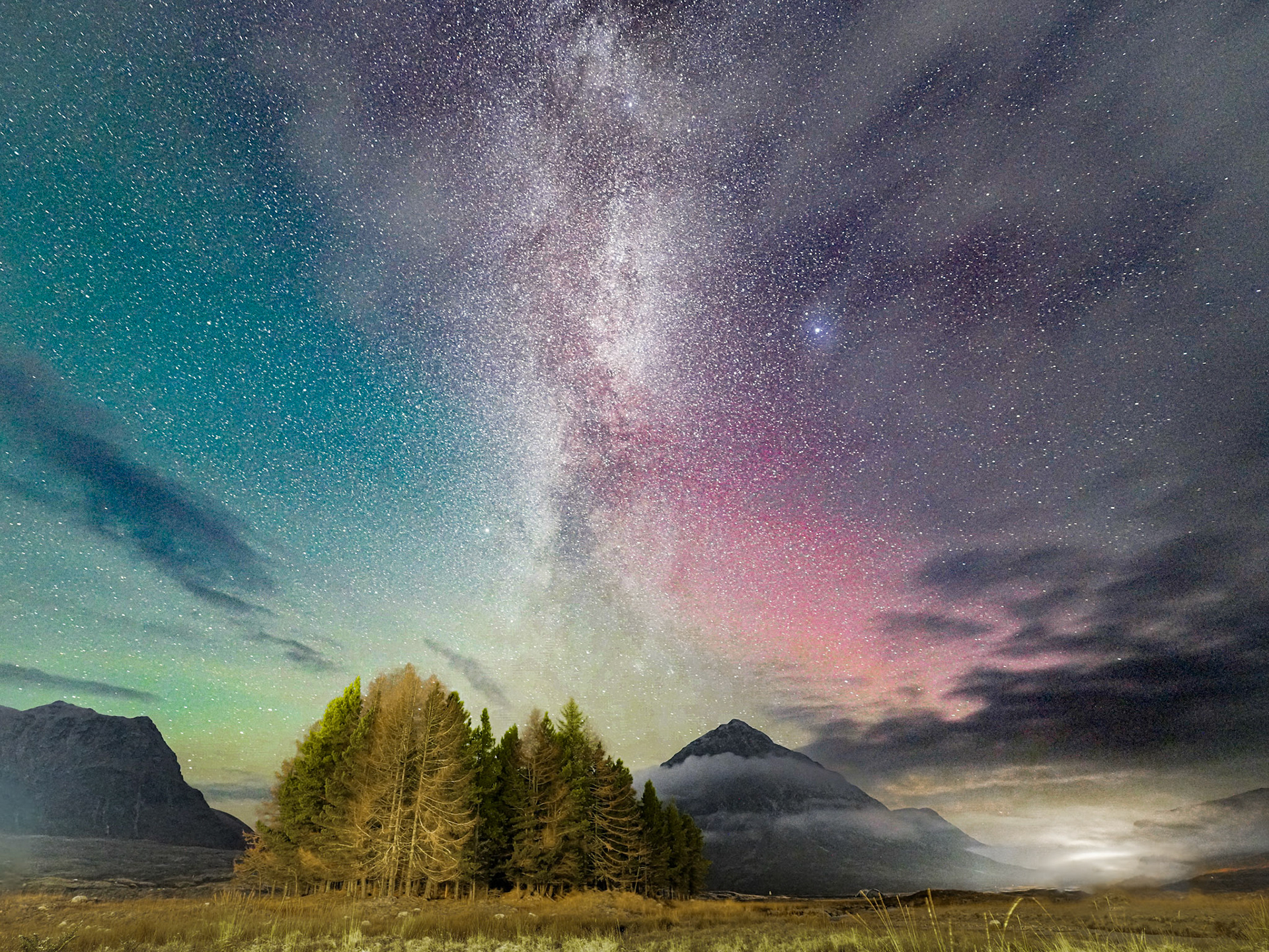 The Milky Way rises vertically above a stand of conifers in a Scottish glen, its core flanked by faint auroral red on the right horizon. Low cloud pools around the base of a pyramidal peak, while green airglow tints the left sky. The coincidence of galactic core, aurora, and valley mist in a single frame is rare and well-judged.