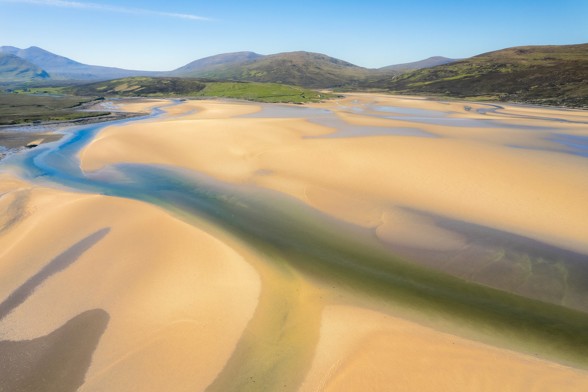 Exposed sandbars and braided tidal channels at low water on a Scottish sea loch, shot from directly above in bright spring light. The sinuous waterways carve abstract forms across the sand, with the mountains of Sutherland receding into haze behind. The aerial vantage reduces the estuary to pattern and colour, balancing graphic structure with a sense of vast, unpeopled space.