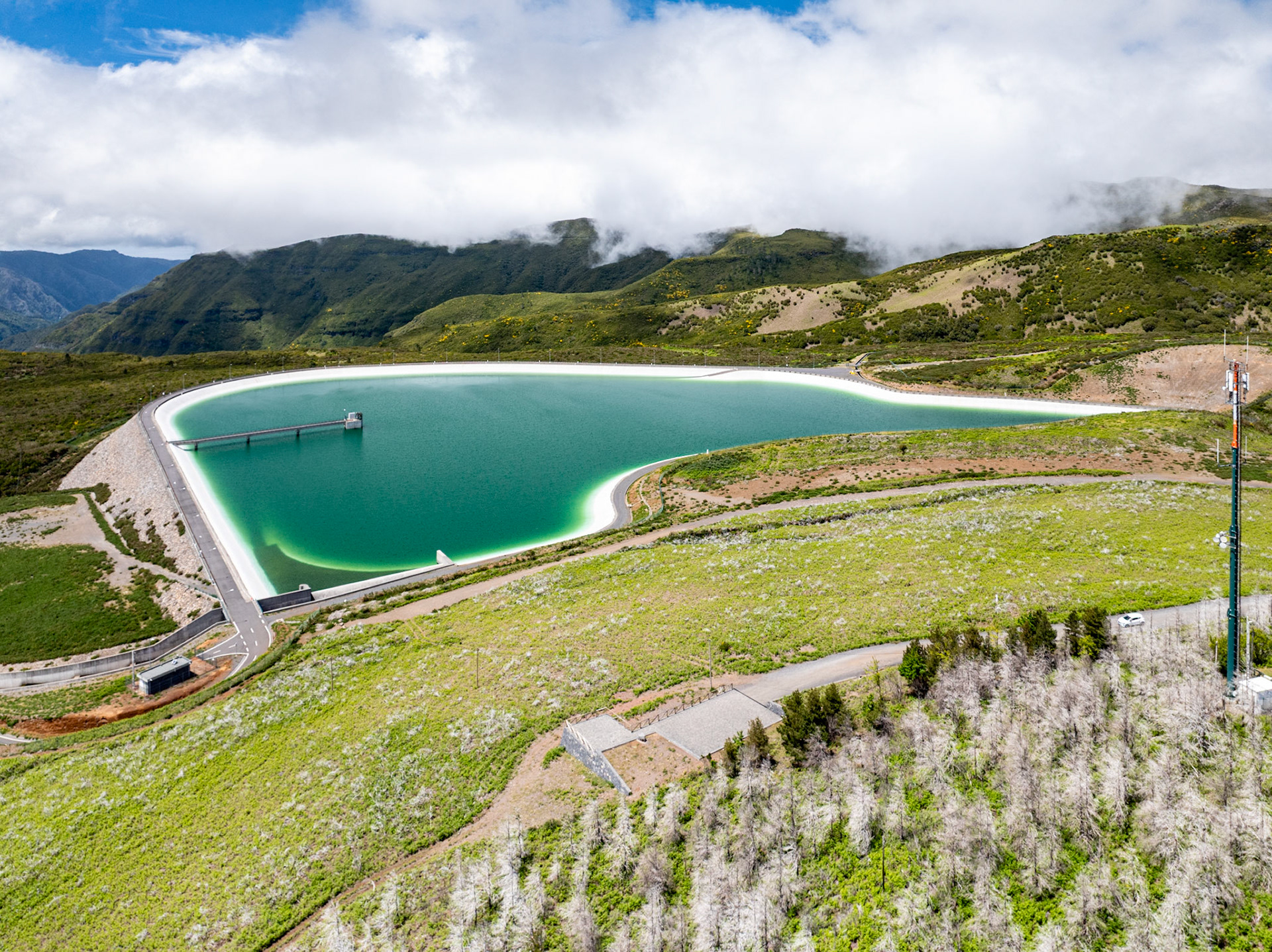 A pumped-storage reservoir sits high above Arco da Calheta on Madeira's southern ridge, its white concrete liner tracing a precise curve against the turquoise water. Low cloud presses against the forested mountains beyond, softening the backdrop without quite lifting into drama. The geometry of infrastructure against the green hillside is the image's only real tension.