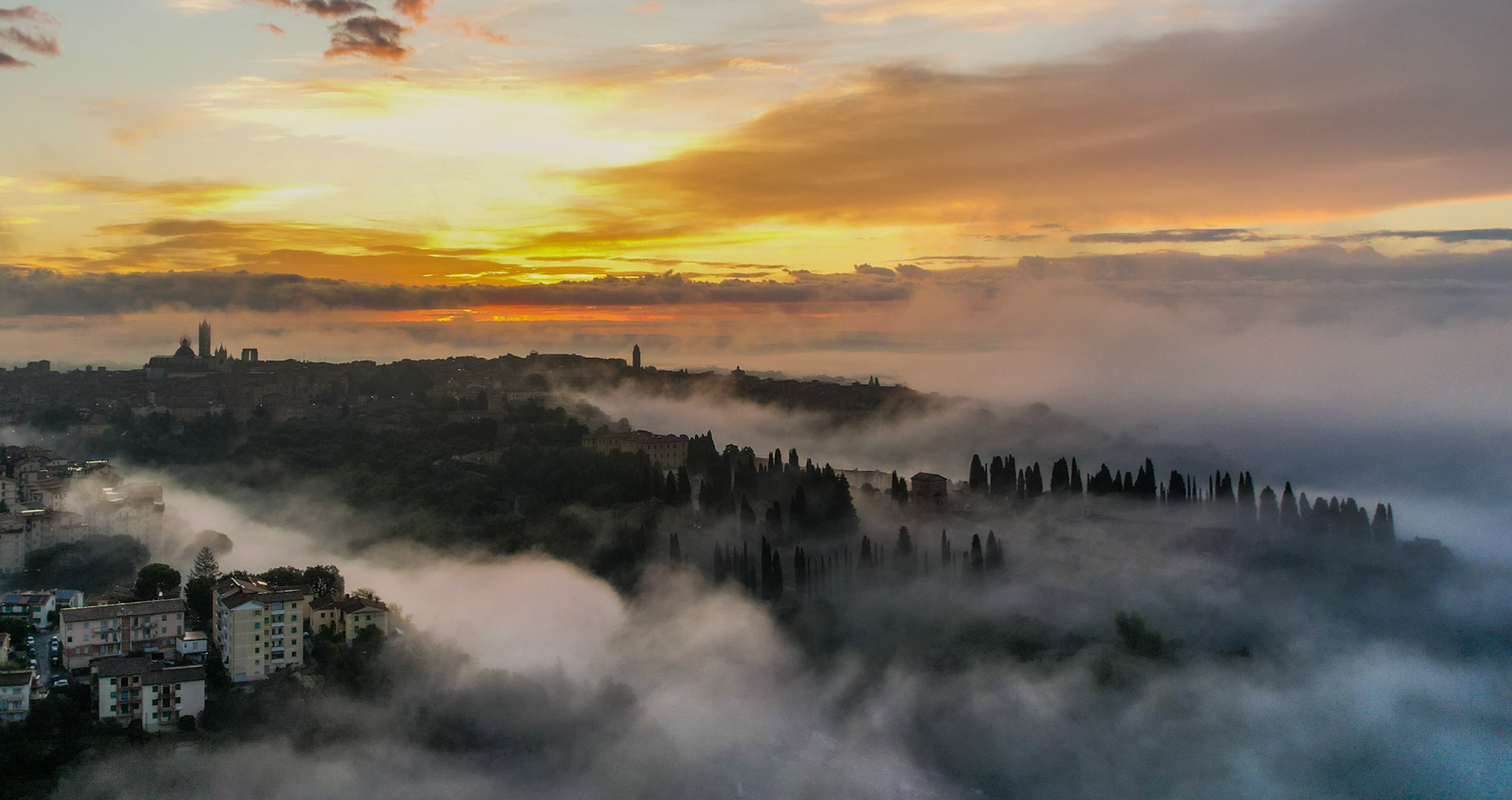 Low inversion fog pools between the hills surrounding Siena at dawn, partially submerging a long avenue of cypress trees and the outlying residential streets. The medieval skyline, including the cathedral silhouette, rises clear above the mist line as the first warm light breaks behind a broken cloud layer. The layering of fog, tree forms, and cityscape gives the frame unusual structural depth for an aerial record shot.