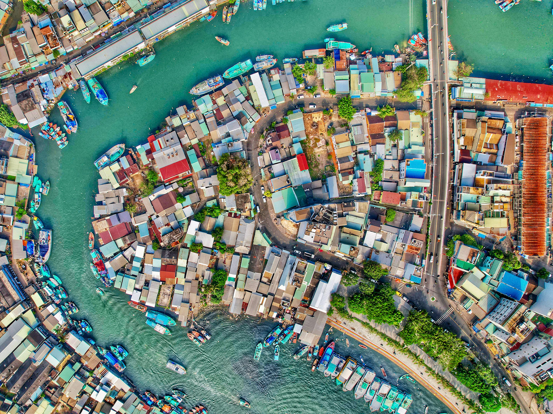 A tidal channel cuts through the dense roofscape of a Vietnamese fishing settlement, its turquoise water threading between corrugated rooftops and moored fishing boats. The nadir perspective flattens the scene into a mosaic of colour and form, the organic sprawl of the village contrasting with the rigid road grid to the east. Captured over Phú Quốc island in January 2023.