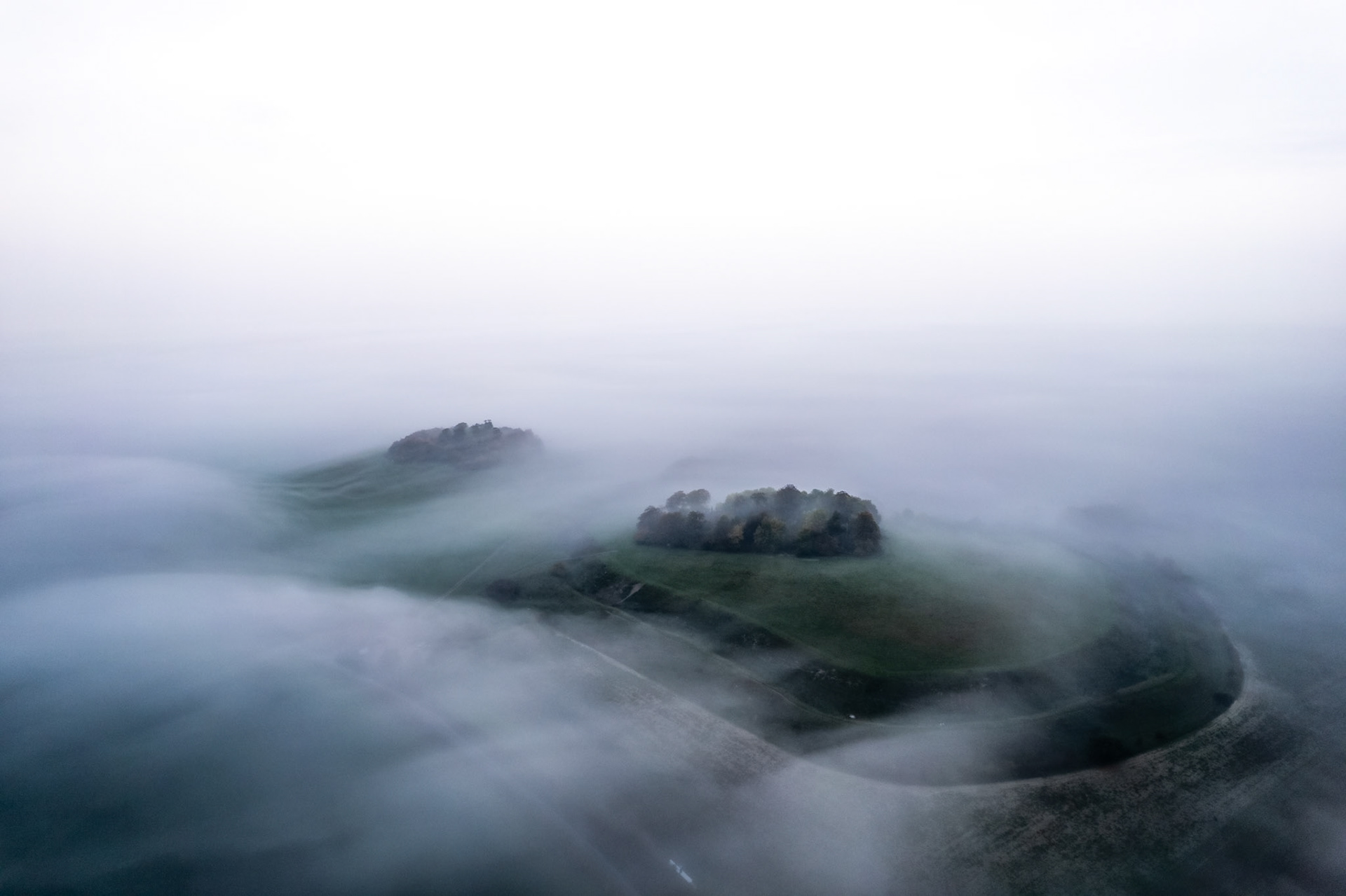 A temperature inversion smothers the Vale of the White Horse, leaving Brightwell Barrow and the Wittenham Clumps in Oxfordshire adrift above a sea of low cloud. The nearer summit retains a crown of trees and a glimpse of road, anchoring the image in landscape while the fog strips everything else to form. Captured by drone at first light on an October morning.