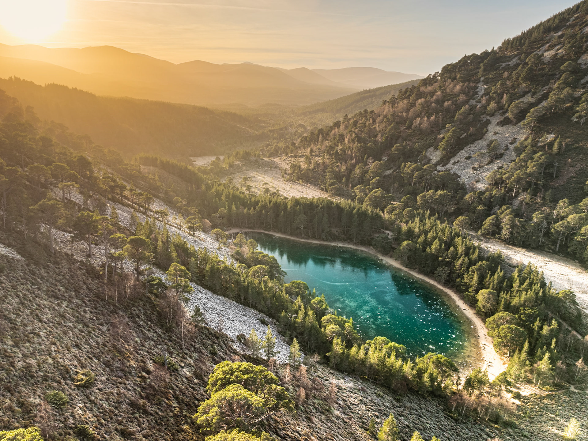 A small lochan catches the last of the winter sun in a pine-forested glen of the Cairngorms, Aviemore. The warm backlight rakes across Scots pine and scree slopes, while layered ridgelines recede into haze. The turquoise water holds its colour against the prevailing amber of the scene.