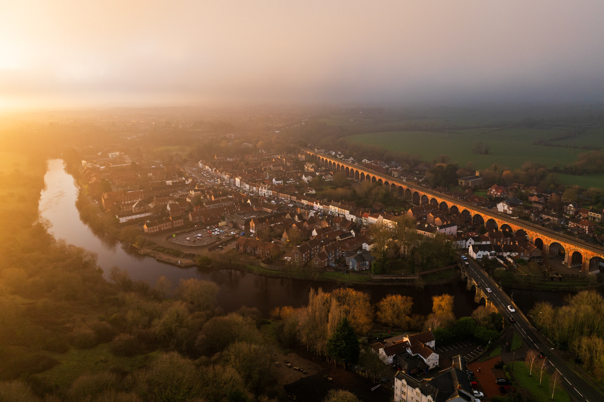 A Victorian railway viaduct bisects a riverside market town as low winter fog softens the horizon and golden light rakes in from the west. The meander of the River Tees anchors the left frame, contrasting the geometric repetition of the arches. Captured at dawn from altitude, the image holds a tension between industrial heritage and quiet atmospheric dissolution.