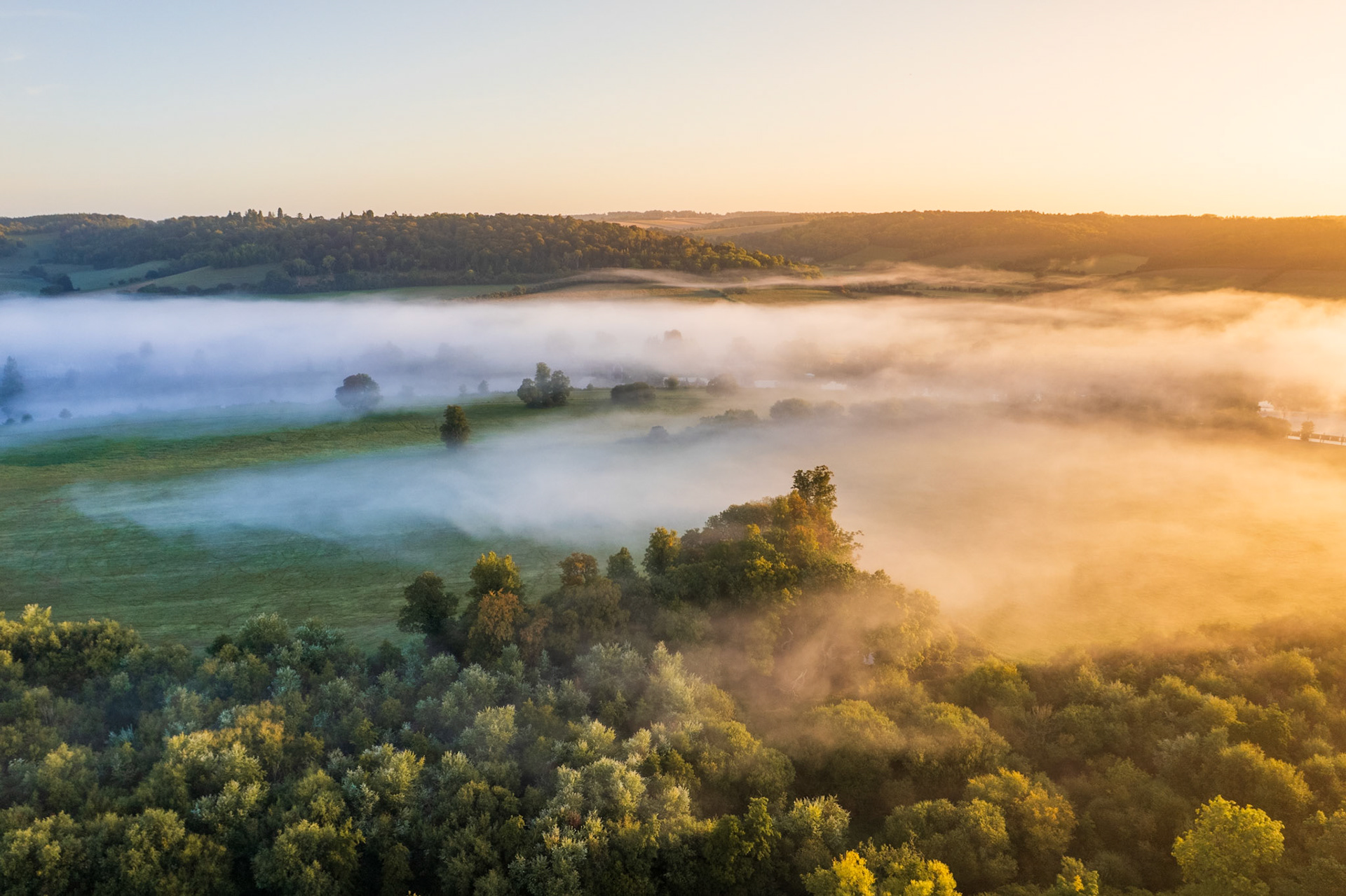 Ground mist pools across a pastoral valley near Henley-on-Thames at dawn, the low sun catching the fog from the south-east and dividing the frame between cool shadow and warm amber light. Deciduous woodland on the valley sides emerges above the mist layer, its canopy already carrying the first tones of early autumn. The aerial perspective reveals the topography beneath the fog in a way impossible from ground level.