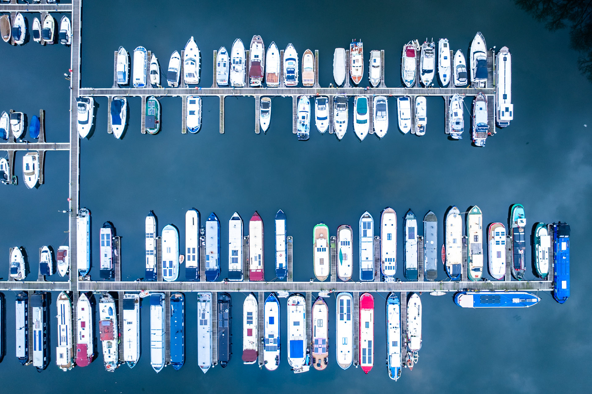 Rows of leisure craft and narrowboats moored in a Thames-side marina near Reading, viewed from directly overhead. The strict geometry of the pontoons and the variety of hull colours create a flattened, almost typographic pattern against the still grey water. Captured in early March, the image reads as urban-industrial inventory rather than landscape.