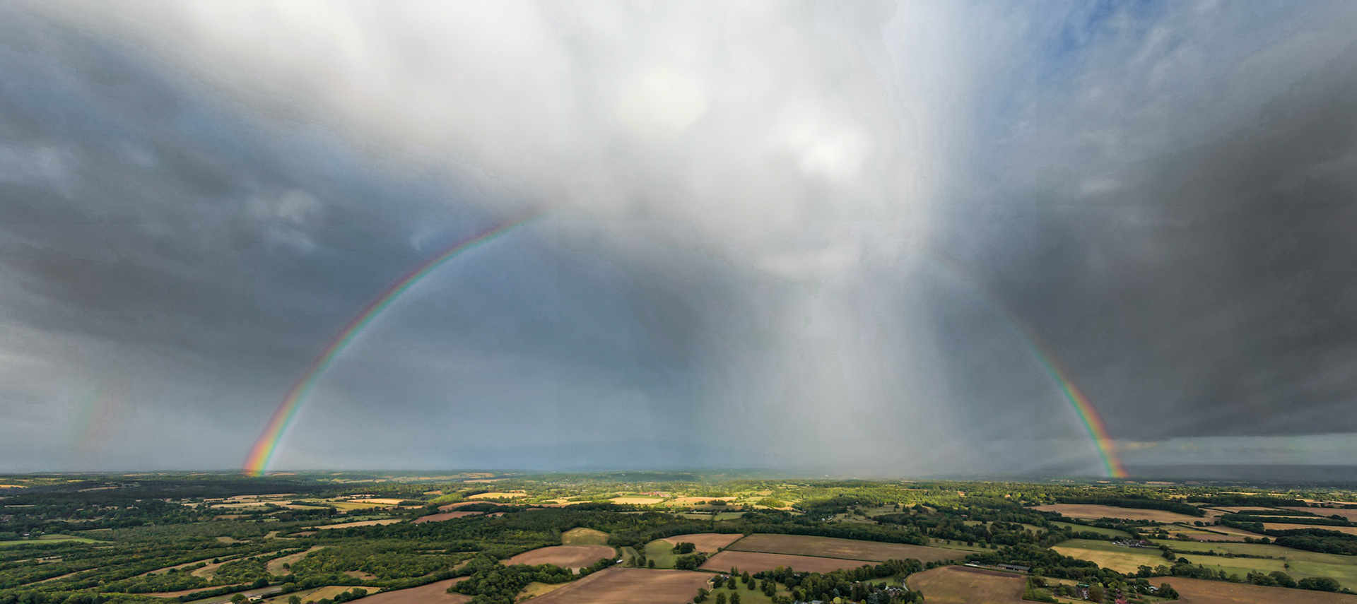 A full primary rainbow spans the patchwork farmland south of Henley-on-Thames, photographed from altitude as a convective storm cell trails virga across the mid-distance. The elevated vantage reveals the complete geometry of the bow against a bruised sky, with sunlit fields caught beneath the arch in sharp contrast to the advancing rain curtain.