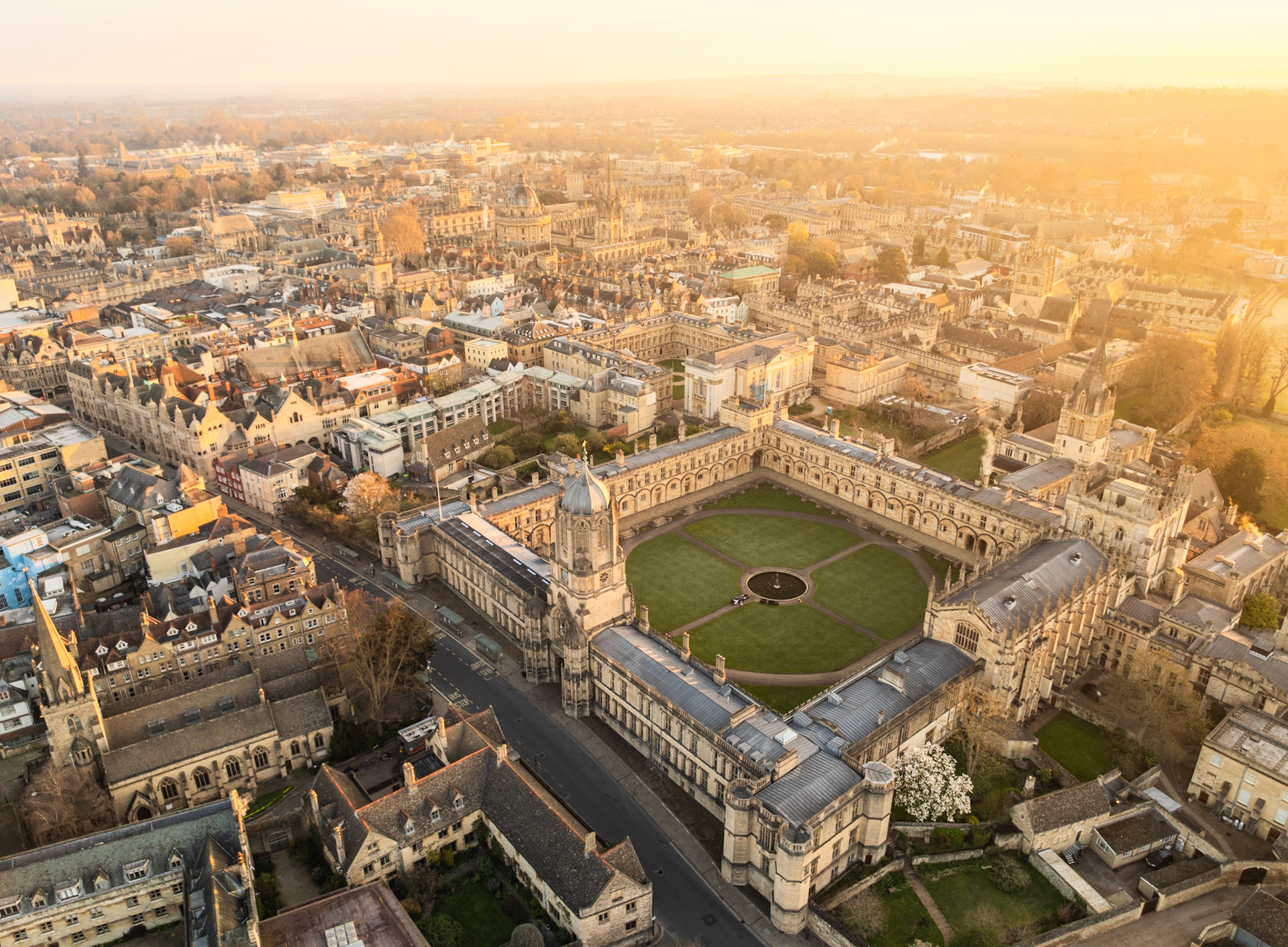 Christ Church College seen from altitude at late-afternoon golden hour in early spring, its quadrangle lawn and central fountain sharply resolved against the honey-coloured limestone of Oxford's medieval core. The warm haze softens the city's edges without obscuring the collegiate geometry that defines the foreground.