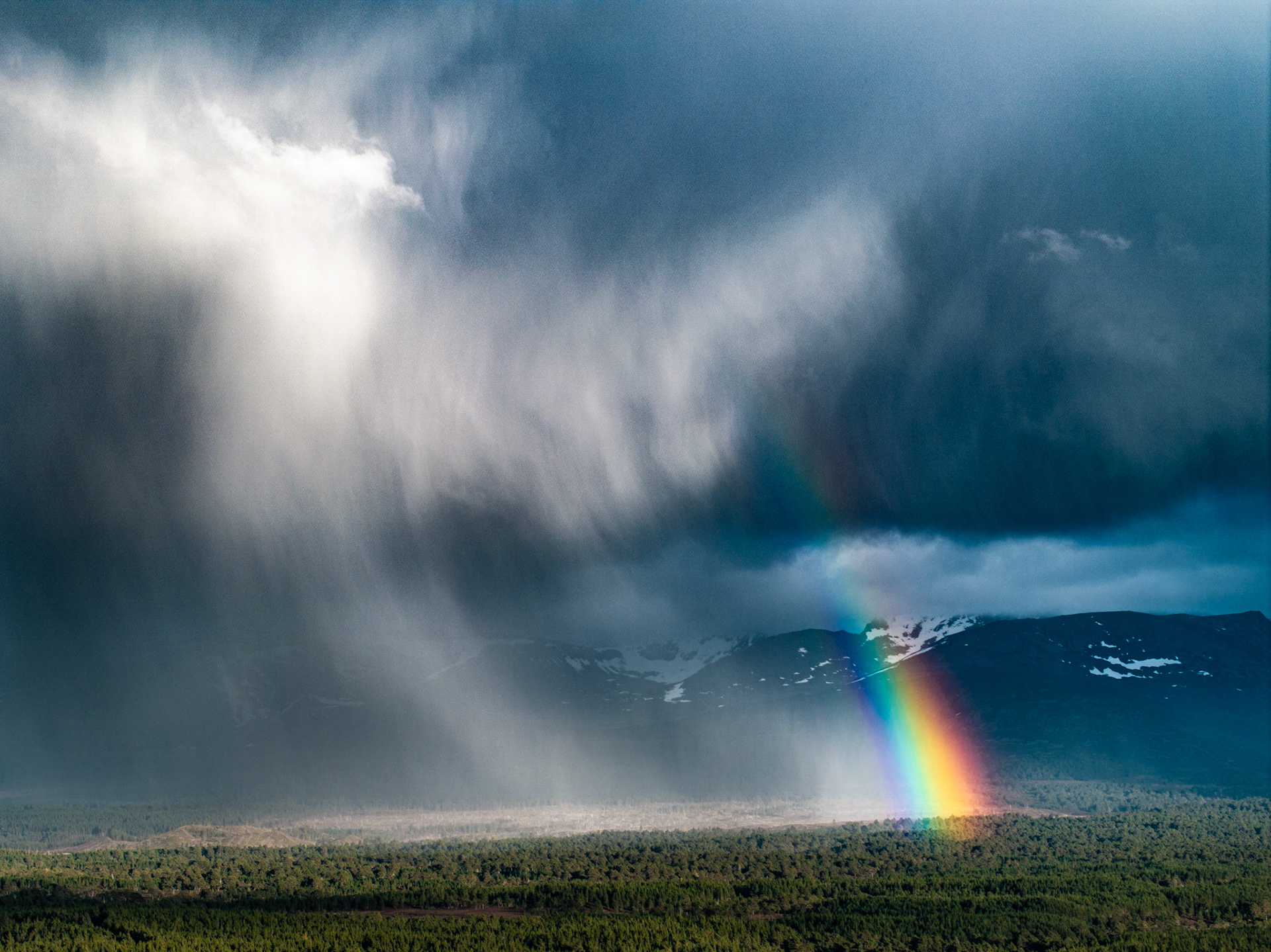 A virga curtain sweeps across the Cairngorm plateau as a vivid rainbow grounds itself at the forest edge near Aviemore. The juxtaposition of precipitation shaft, snow-patched summit, and saturated spectral arc within a single frame captures a fleeting atmospheric collision rarely framed with such compositional weight. Aerial elevation compresses the layers — boreal forest, moorland, mountain and sky — into a single dramatic plane.