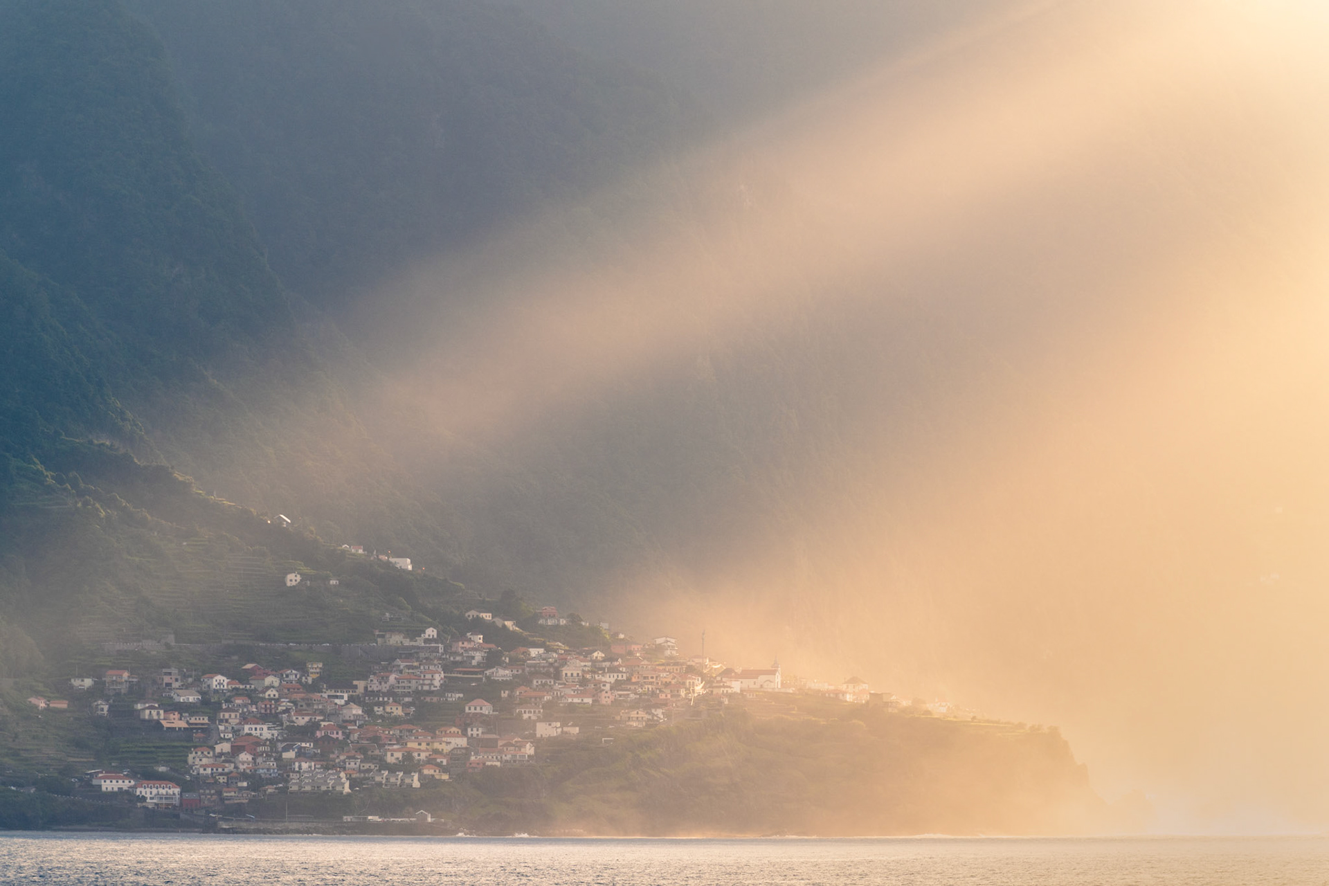A coastal settlement clings to steep terraced slopes as a wall of mist rolls in from the sea, splitting the scene between cool shadow and warm evening light. The village — most likely on Madeira's north coast — is caught at the threshold of erasure, the golden haze reducing white-walled houses to silhouette at its margin.