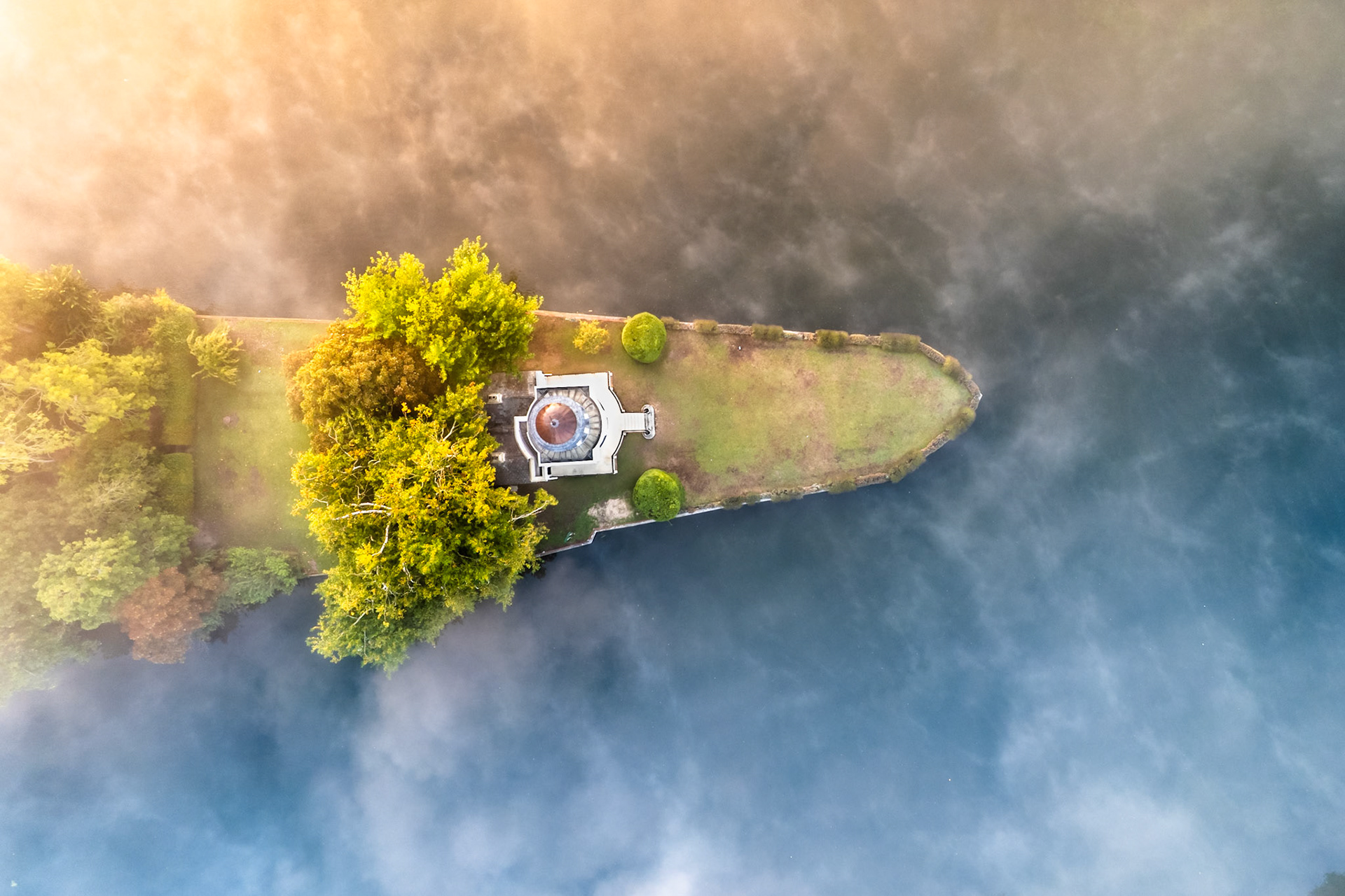 A narrow island on the Thames near Henley emerges from low dawn fog, its domed folly and manicured lawns sharp against the mist-blurred water. Morning light breaks from the upper left, catching the tree canopy and casting a warm contrast against the cool grey vapour that diffuses the river surface. The aerial perspective reduces the scene to a near-abstract arrangement of landform and atmosphere.
