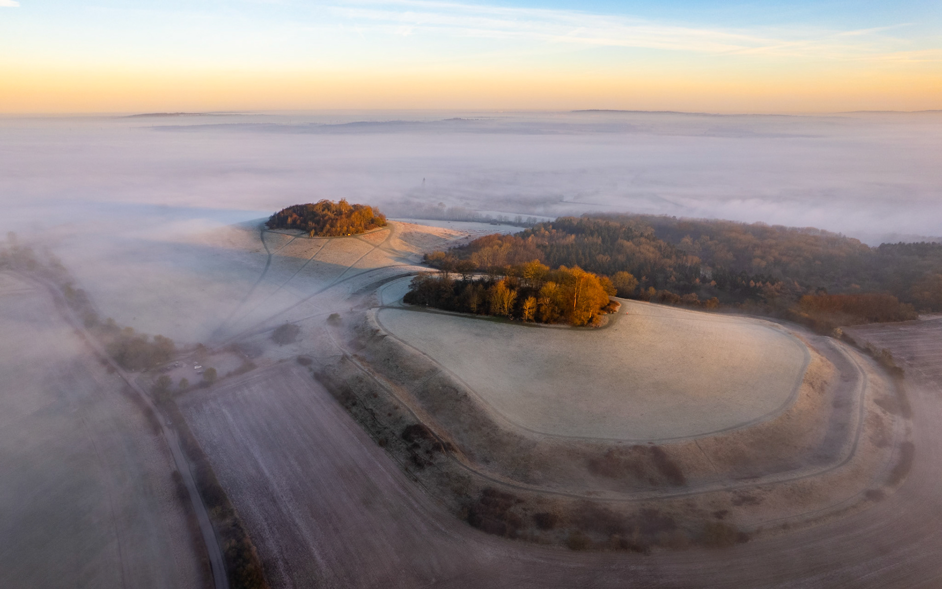 A temperature inversion buries the Vale of White Horse beneath a still sea of fog, viewed from Brightwell Barrow near the Wittenham Clumps in Oxfordshire, leaving frost-covered hilltops and copses of bare-branched trees standing clear in early dawn light. The curving field margins and converging ridge lines give the elevated ground an almost archipelago quality. Warm tones from the low sun catch the crowns of the remaining autumn-holdover foliage against the cold grey below.