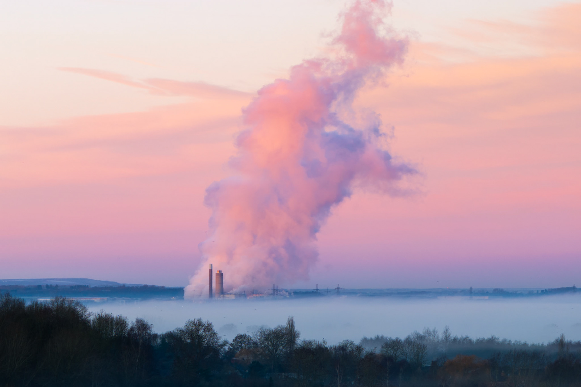 A temperature inversion locks ground fog across the Thames Valley near Wallingford at dawn, while steam and smoke from a distant power station billow upward into a pink and violet afterglow sky. The chimneys barely clear the fog layer, the plume dwarfing the industrial structures beneath it. Shot from a drone in January 2026, the image turns an environmental subject into an exercise in tone and scale.