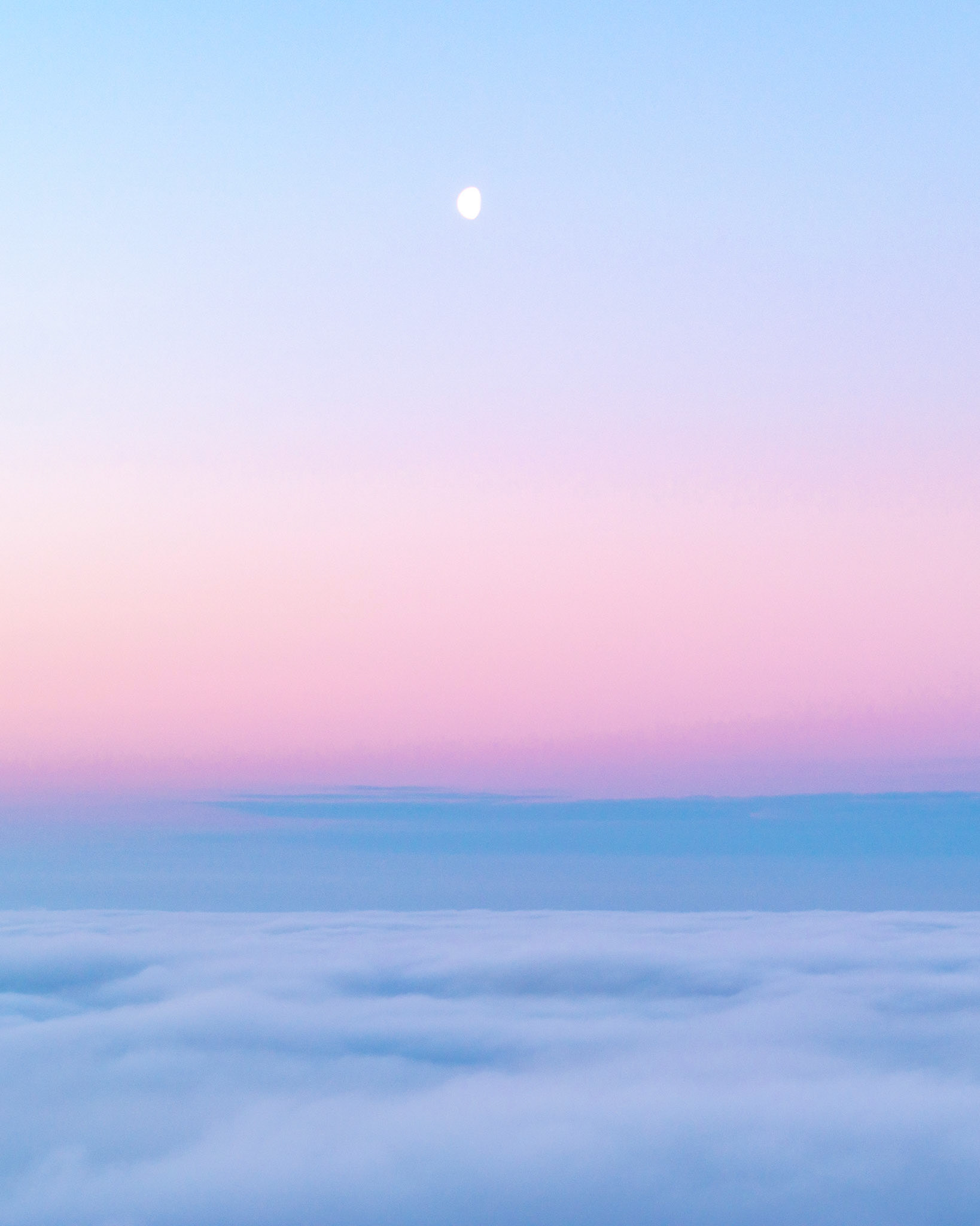 A temperature inversion buries the Thames Valley beneath a flat sea of cloud at dusk, the gibbous moon suspended in a sky grading from powder blue through rose to lavender. Shot from elevation above Abingdon-on-Thames, the compression between cloud ceiling and clear upper atmosphere creates a horizon line of unusual stillness. The near-total absence of hard edges gives the frame a quality closer to watercolour than photography.
