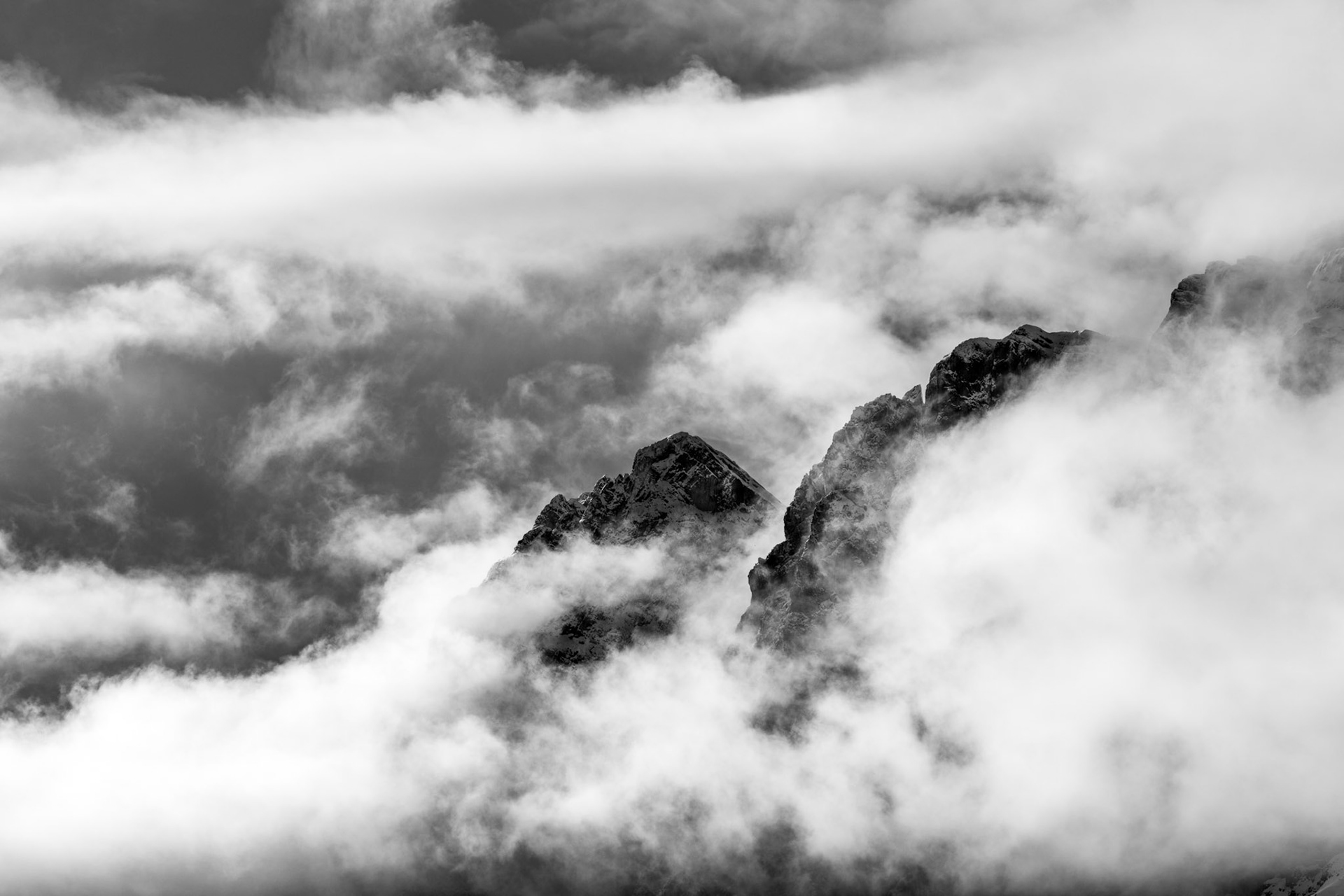 Rocky summits pierce a churning sea of cloud, their dark ridgelines rendered in sharp contrast against swirling mist. The image compresses depth so that mountain and weather become almost equal subjects, the rock asserting itself briefly before cloud closes again. Captured in mid-March, residual winter conditions cling to the upper faces.