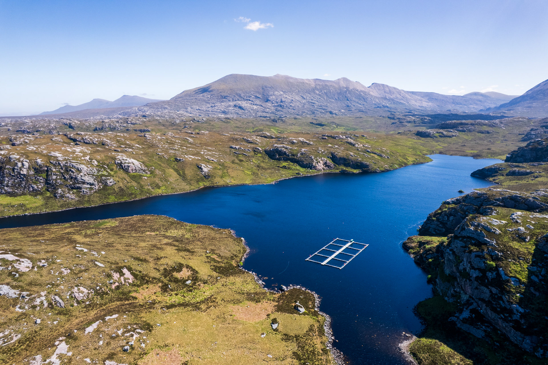 A fish farm installation sits on a dark Highland loch amid quartzite moorland north of Lairg, Sutherland. The industrial structure anchors the eye but pulls against the wilderness scale of the scene. Midday light flattens the terrain and removes the atmospheric separation that might otherwise elevate the composition.