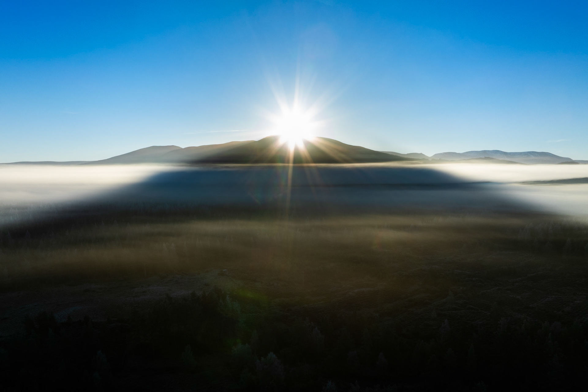 A temperature inversion fills the Strathspey valley floor as the sun crests a hill above Aviemore, casting the mountain's shadow across the fog layer in a near-symmetrical wedge. Captured from altitude on a clear December dawn in the Cairngorms, the image records the brief moment when the shadow geometry aligns with the mist boundary. Lens flare from direct sun contact adds an unplanned graphic element that competes with the composition.