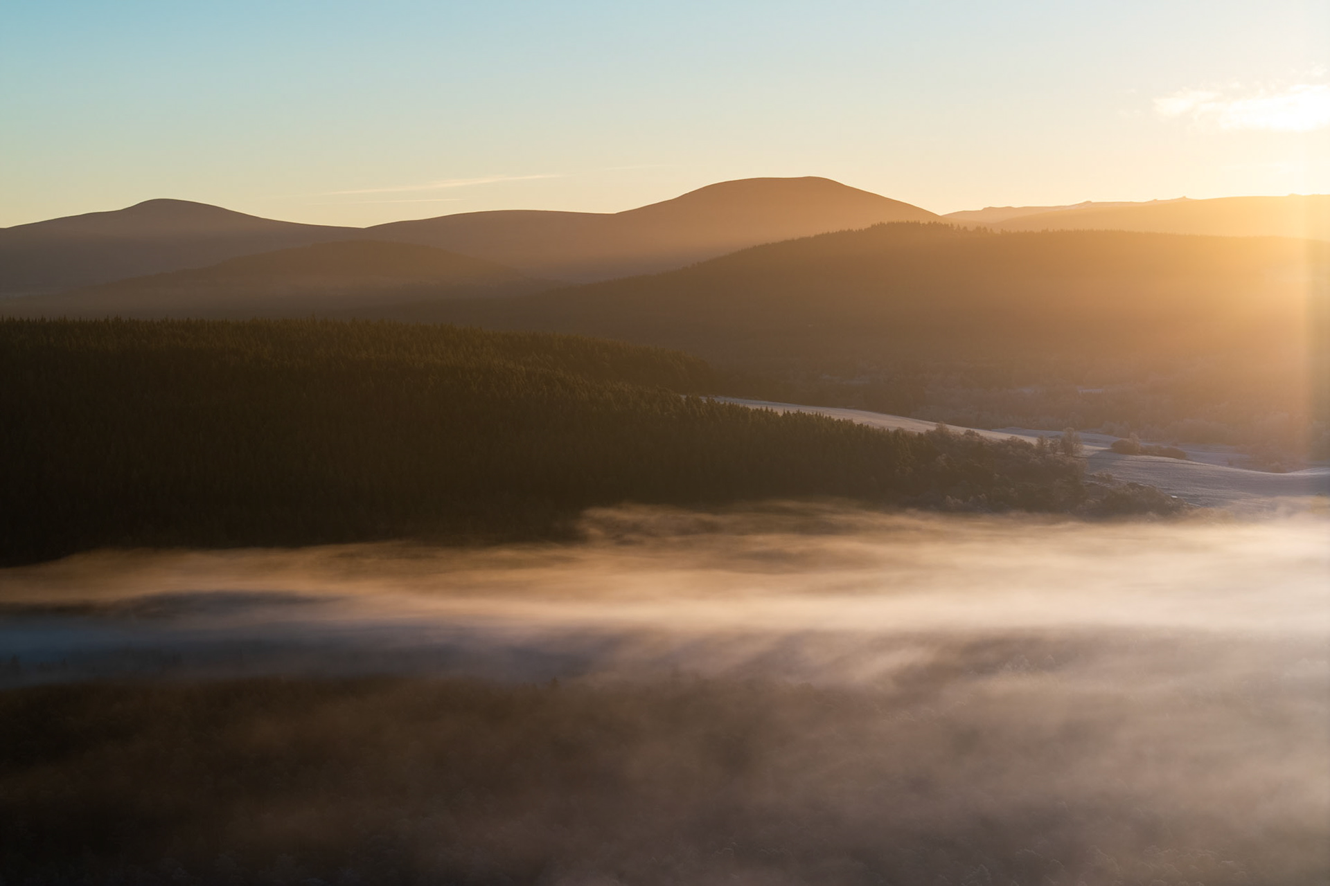 A temperature inversion fills the Nethy Bridge valley with low mist on a winter solstice dawn, the Cairngorm hills receding in warm silhouette against a clear sky. The tree line of Abernethy Forest separates fog from mountain, compressed into distinct tonal bands by the low sun entering from the right. A river channel threads briefly into view before disappearing beneath the cloud.
