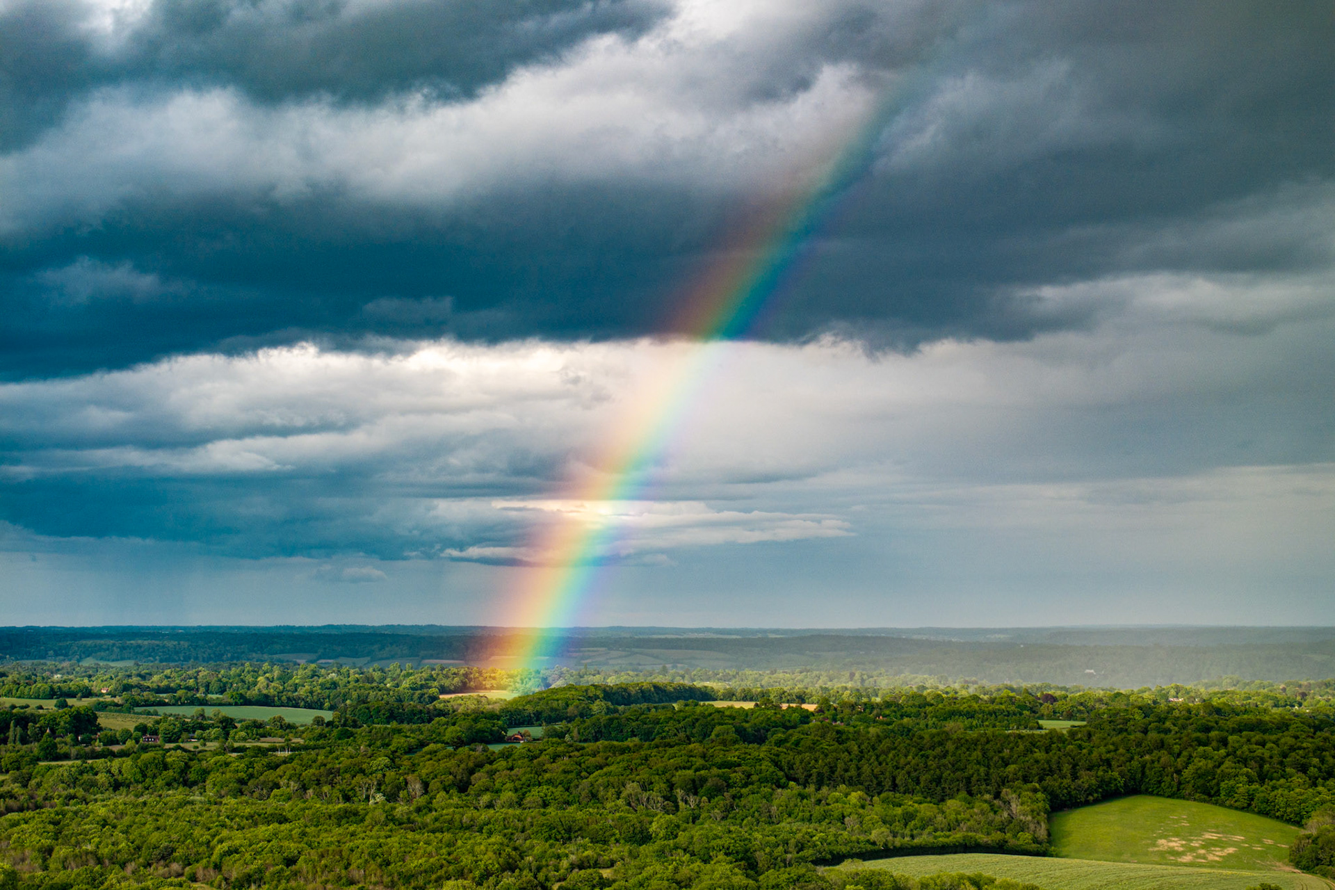 A vivid primary rainbow descends toward the wooded escarpment south of Henley-on-Thames, its foot planted in spring canopy illuminated by a break in passing storm cloud. Shot from altitude, the aerial perspective compresses woodland, farmland and rain curtain into a single dramatic plane. The saturation of the arc against the dark cumulonimbus is entirely natural, owing nothing to post-processing excess.
