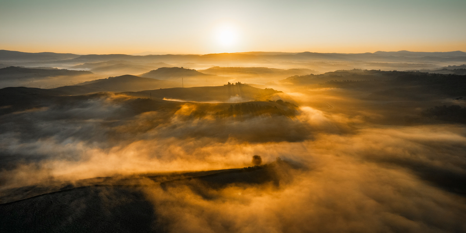 Low-lying fog pools between the rolling hills of the Crete Senesi at dawn, lit from behind by a rising sun still close to the horizon. A lone farmstead on an isolated hillock breaks the surface, anchoring the composition amid the dissolving mist. The image was captured from altitude, compressing the layered landforms into a quiet, painterly sequence of receding planes.