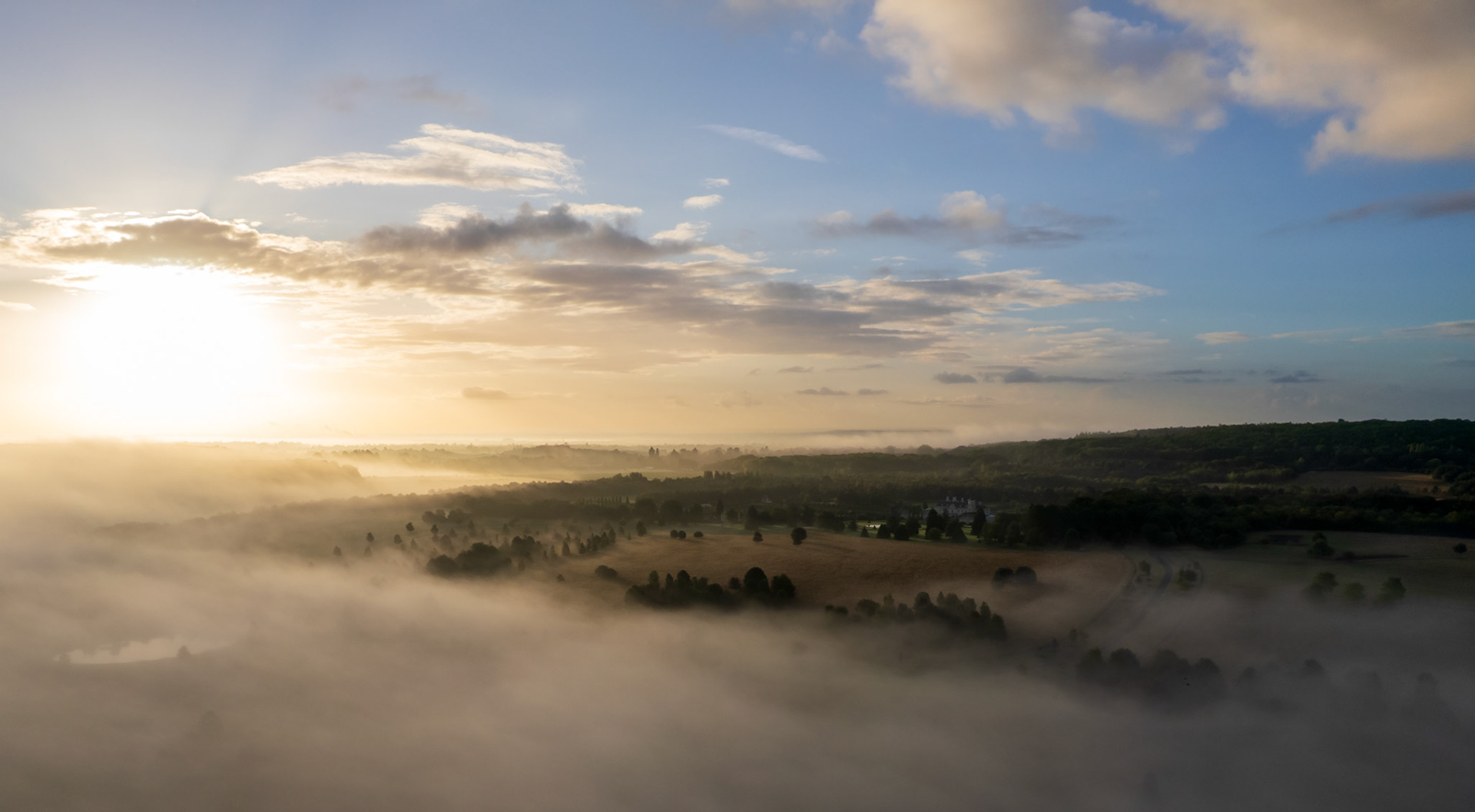 A temperature inversion holds a blanket of fog across the Thames Valley lowlands near Maidenhead, photographed from altitude at first light on an early September morning. Isolated tree crowns and field boundaries break the fog surface, while warm light from the low sun diffuses through the moisture-laden air to the west. A country house sits half-revealed in the middle distance, anchoring the otherwise abstract pastoral scene.