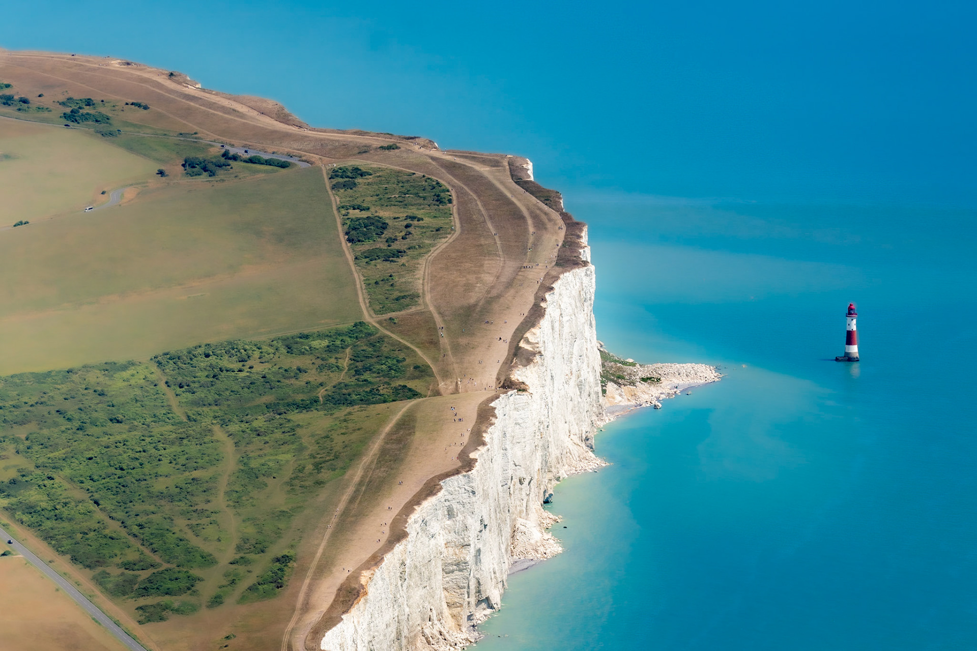 Beachy Head's chalk cliffs fall sheer to a flat calm English Channel, the red-and-white banded lighthouse standing isolated on its rock platform below. Shot from a high vantage at midday in high summer, the image trades atmosphere for scale and graphic clarity. The parched downland above the cliff face records the dry conditions of July.