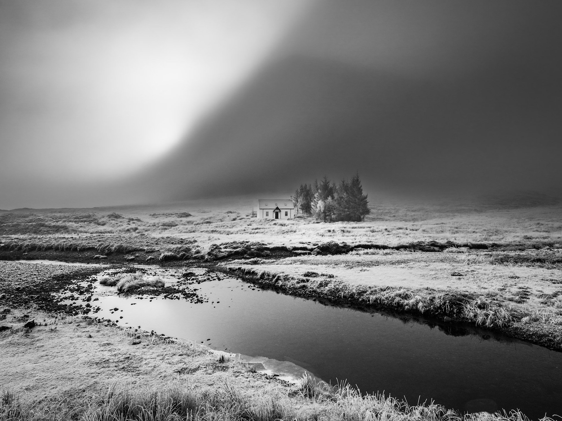 A solitary farmhouse and small stand of conifers sit exposed on a frost-bleached moor as a storm front sweeps overhead, its dark mass bisected by a shaft of diffuse light. A shallow moorland stream curves through the foreground, its still surface reflecting the unsettled sky. The tonal contrast between the luminous cloud break and the advancing darkness gives the image its underlying tension.