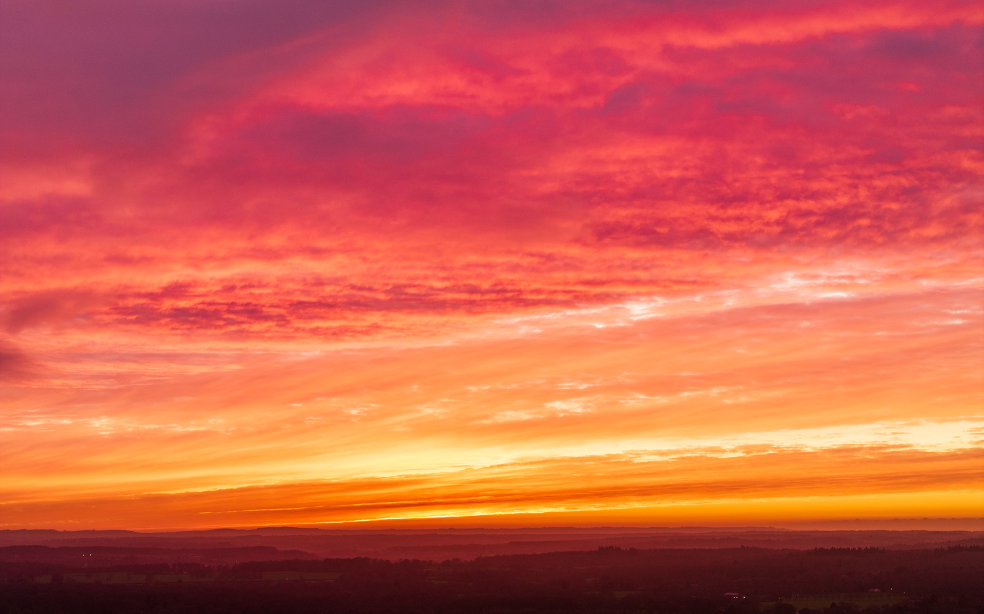 A winter dawn ignites the sky above the Thames Valley, seen from altitude near Henley-on-Thames. Successive cloud layers catch the afterglow in bands of crimson, amber, and gold, while the darkened patchwork of woodland and fields below provides minimal counterpoint. The composition is weighted almost entirely to sky, with little to anchor the foreground.