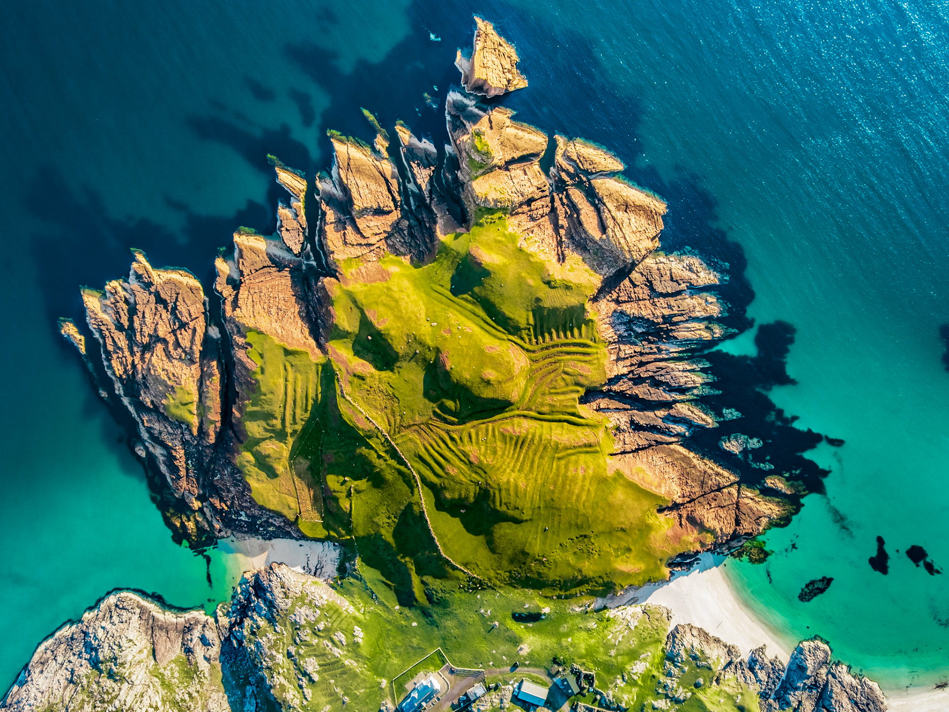 A tidal headland on the north-west Scottish coast, photographed from altitude in late evening light. Folded sedimentary strata radiate outward like fins from a grass-capped central plateau, while ancient lazybeds score the turf in parallel ridges. The surrounding sea shifts from deep indigo to shallow turquoise where sand lies beneath the surface.