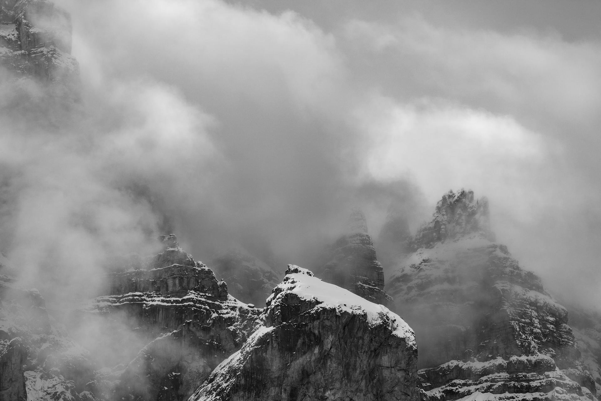 Snow-dusted dolomitic rock towers emerge briefly through churning winter cloud, their layered geology rendered in sharp relief against the diffuse grey above. The monochrome conversion strips all warmth from the scene, leaving only texture and depth as cloud wraps and obscures the upper pinnacles. A moment of partial revelation in otherwise closed, stormy mountain conditions.