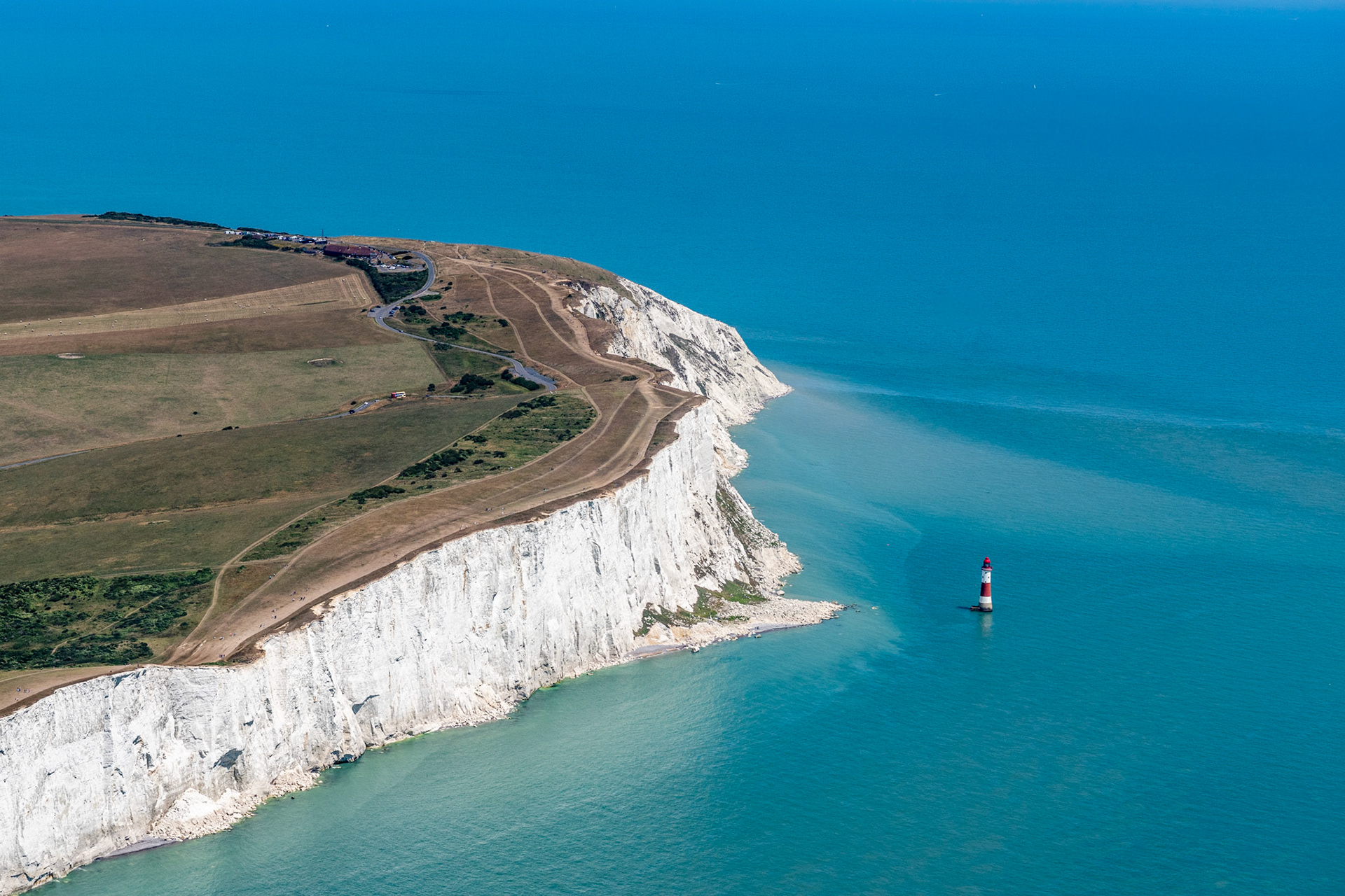 Beachy Head lighthouse stands isolated at the base of the chalk cliffs, dwarfed by the headland's sweeping face. Shot from a high lateral vantage, the image pits the geometry of eroded chalk strata against the flat blue of the English Channel. The midday light is clean but harsh, flattening shadow detail across the cliff face.