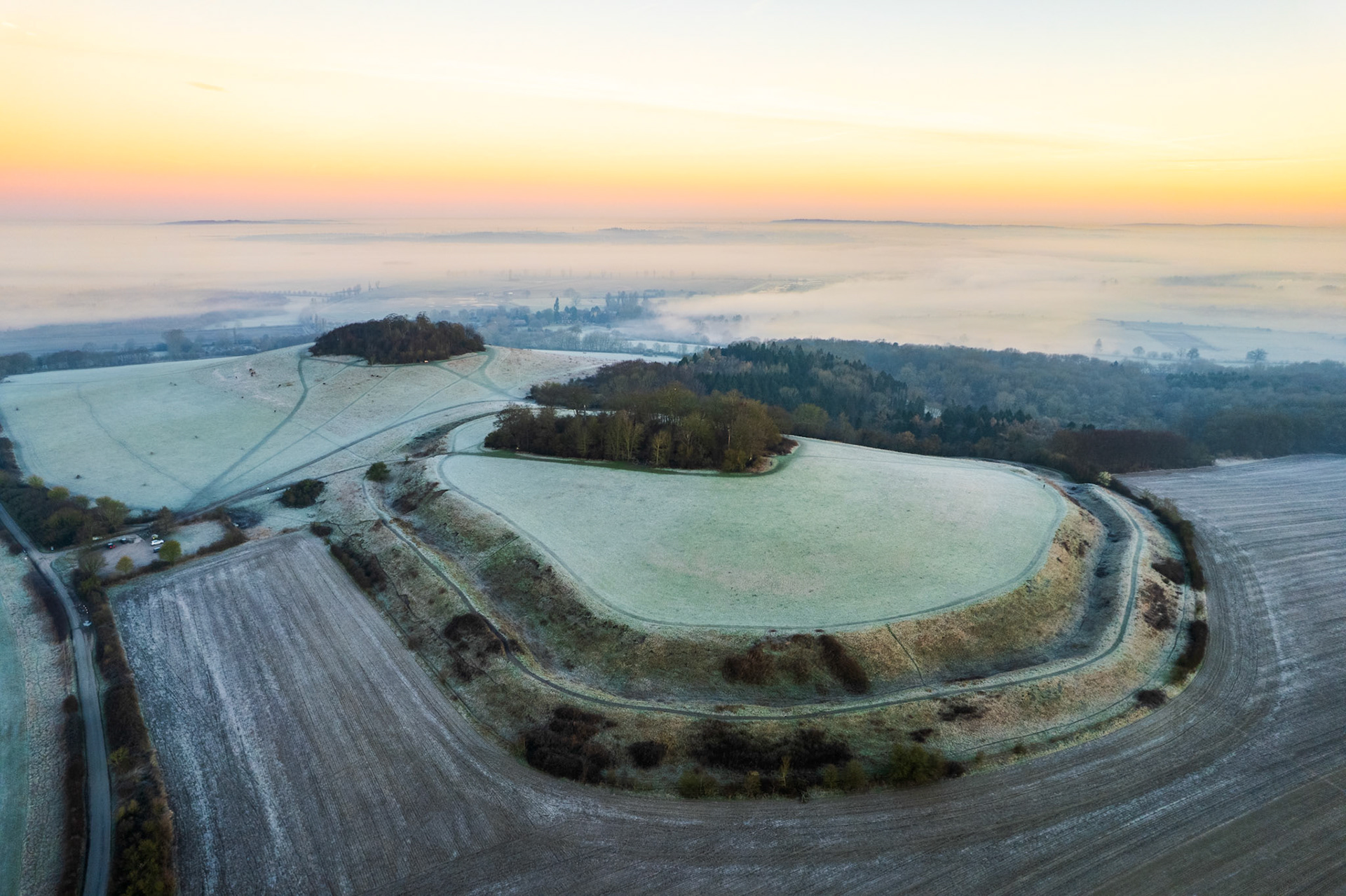 Frost covers the chalk earthworks of Brightwell Barrow, the Iron Age hillfort that sits above the Wittenham Clumps ridge in Oxfordshire, while temperature inversion cloud obscures the valley floor toward Abingdon-on-Thames below. The curved ramparts and field boundaries describe a near-perfect arc against the white-draped lowlands. A warm pre-sunrise horizon holds the moment between mist and open sky.