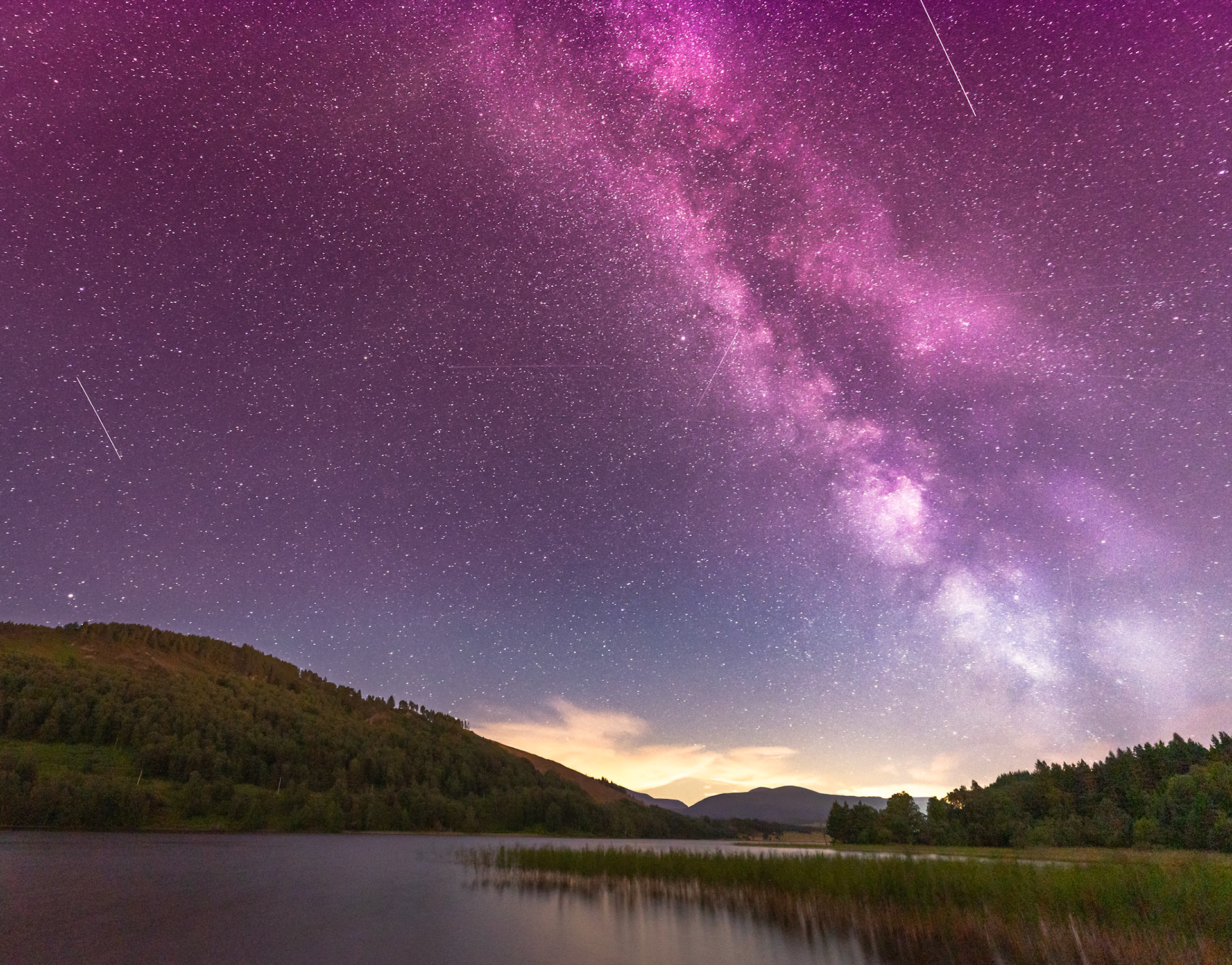 The Milky Way core rises above a wooded Highland loch on a clear August night, its magenta-violet galactic band arching from horizon to zenith. A warm afterglow lingers behind the distant Cairngorm plateau while multiple satellite or meteor trails streak the deep-sky field. Still water in the foreground mirrors the darkness of the scene without resolving a clean reflection.
