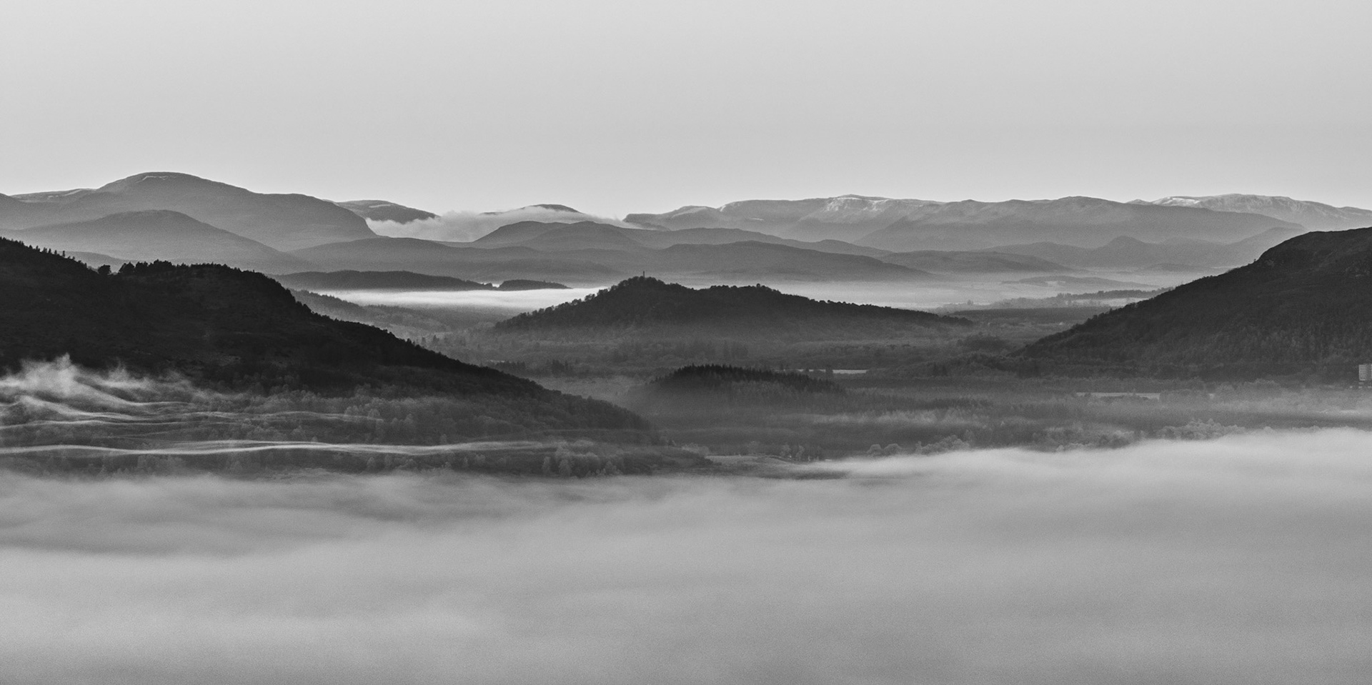 A temperature inversion fills the Spey Valley on Christmas morning, submerging the low ground in dense fog while successive ridgelines recede into haze above Boat of Garten. The monochrome conversion distils the scene to pure tonal recession, with a hilltop structure — possibly a monument — emerging as the sole punctuation in the middle distance. Aerial elevation reveals the full depth of the layering in a way unavailable from the valley floor.